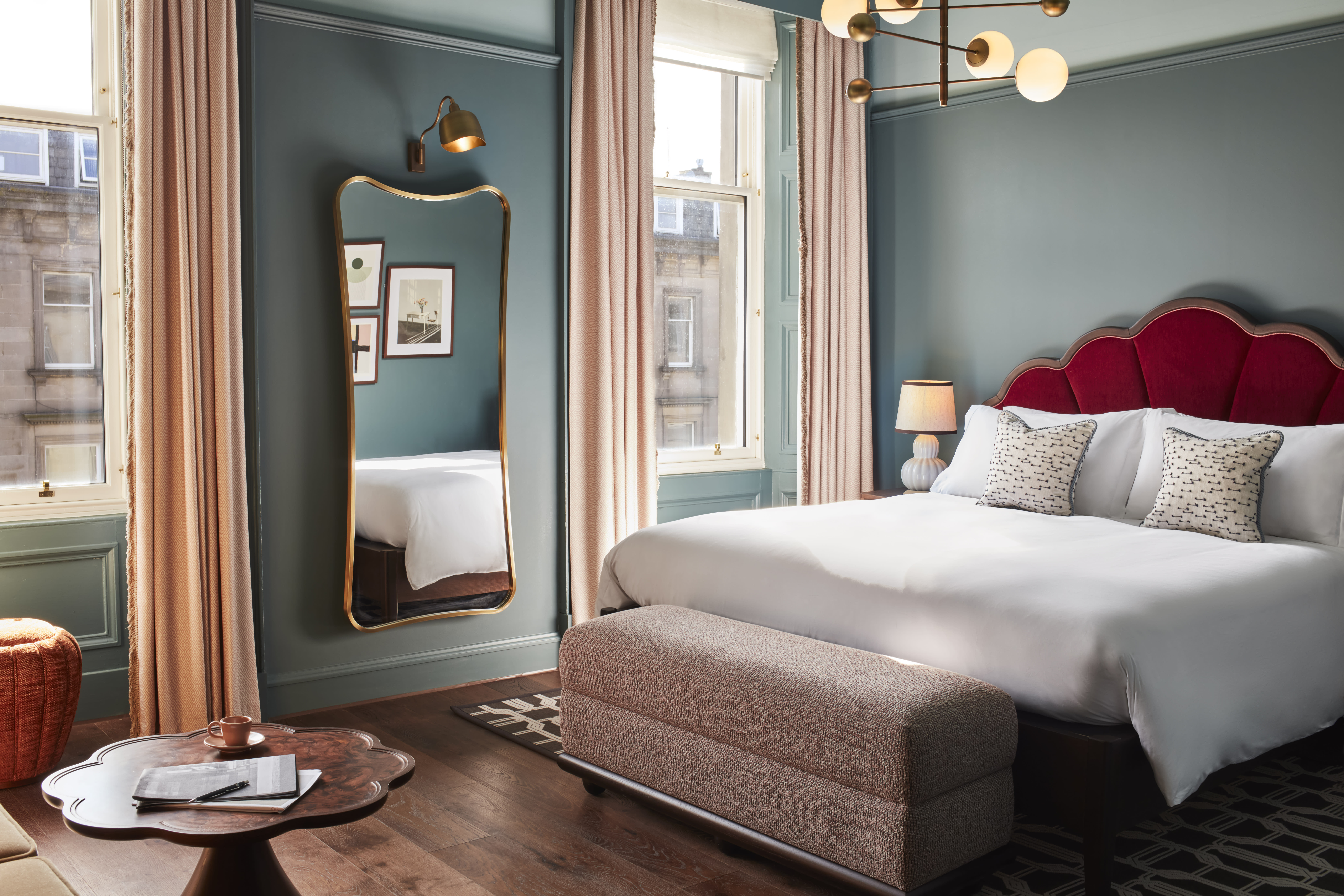 Stylish hotel room with teal walls, a red velvet scalloped headboard, soft pink curtains, and a large gold-framed mirror reflecting natural light from two windows.