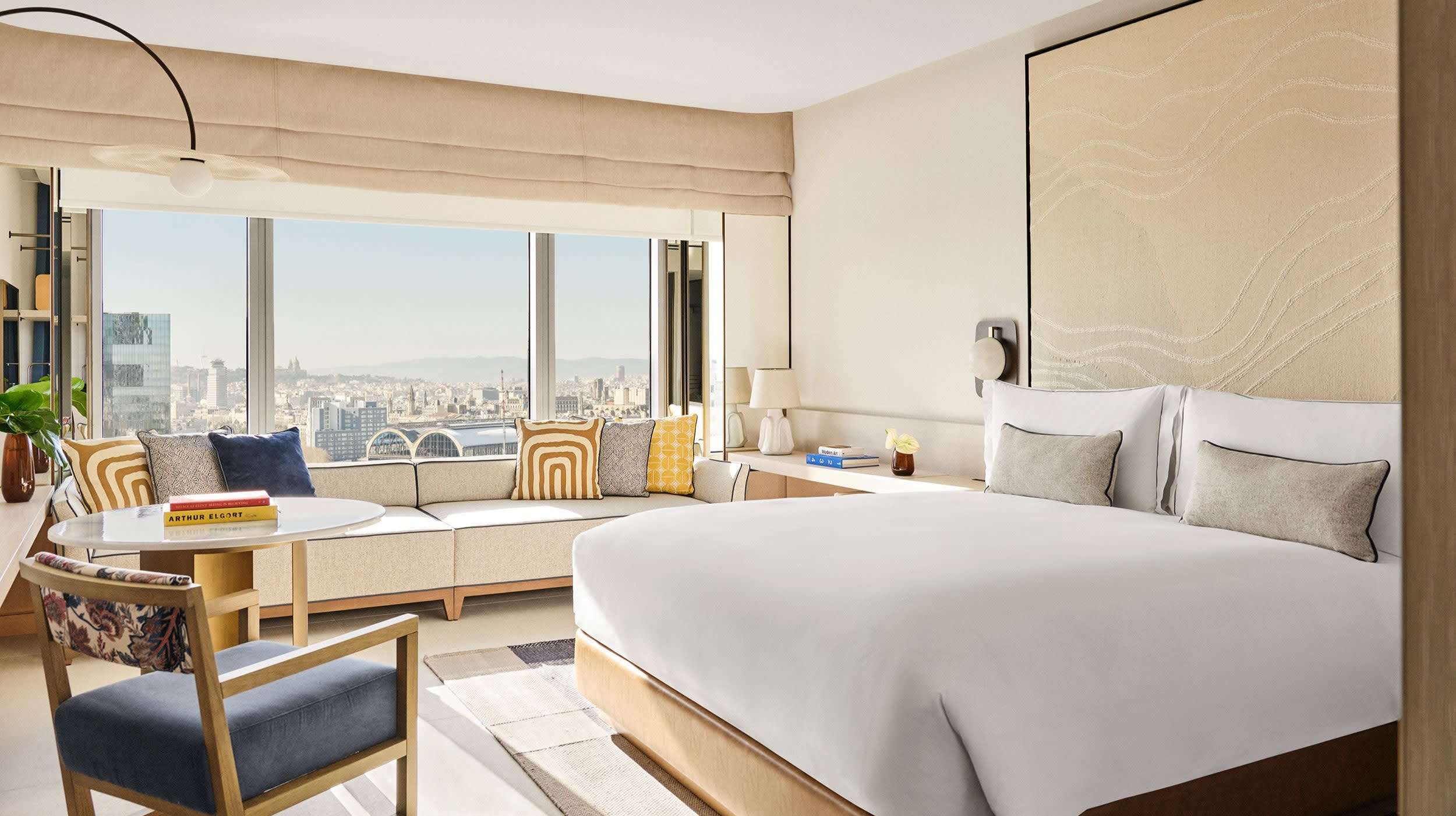 Bright and airy hotel room with a large bed, beige and tan tones, a window-side couch with colorful cushions, and a sweeping city view through floor-to-ceiling windows.