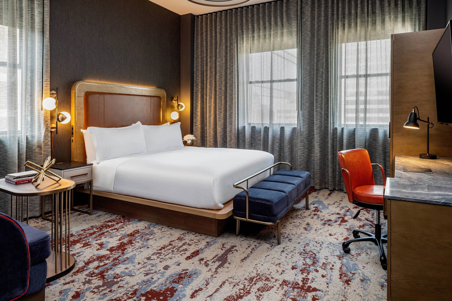 A dark walled hotel room with brown headboard, white linens, blue ottoman and red desk chair