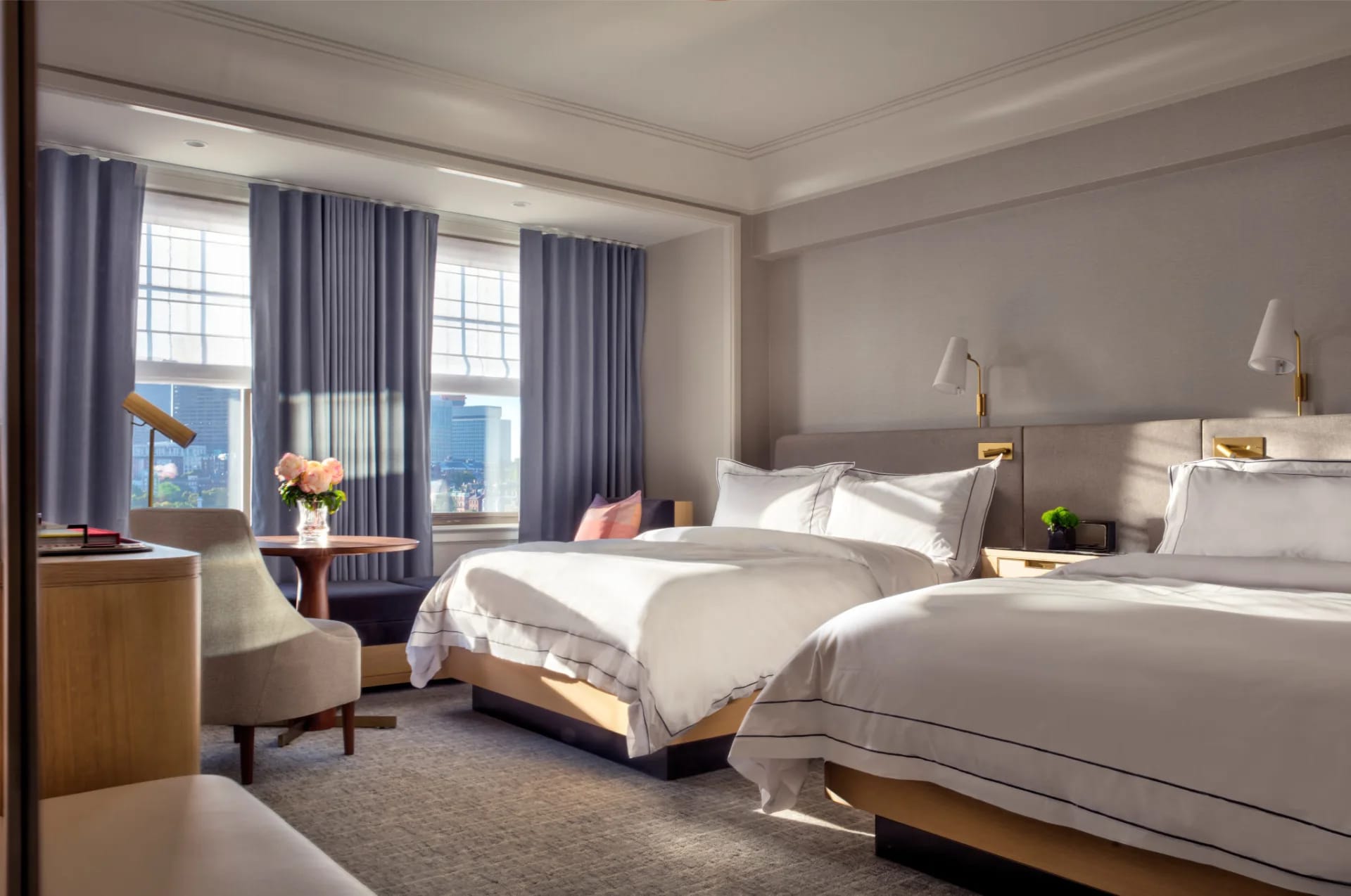 double beds with white linens in a hotel room with light blue curtains and grey chair