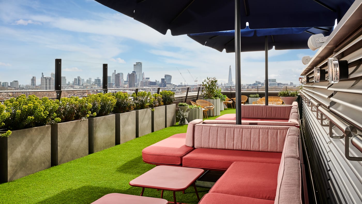 A colorful rooftop terrace with pink cushions and lounge furniture and green synthetic grass overlooking London.