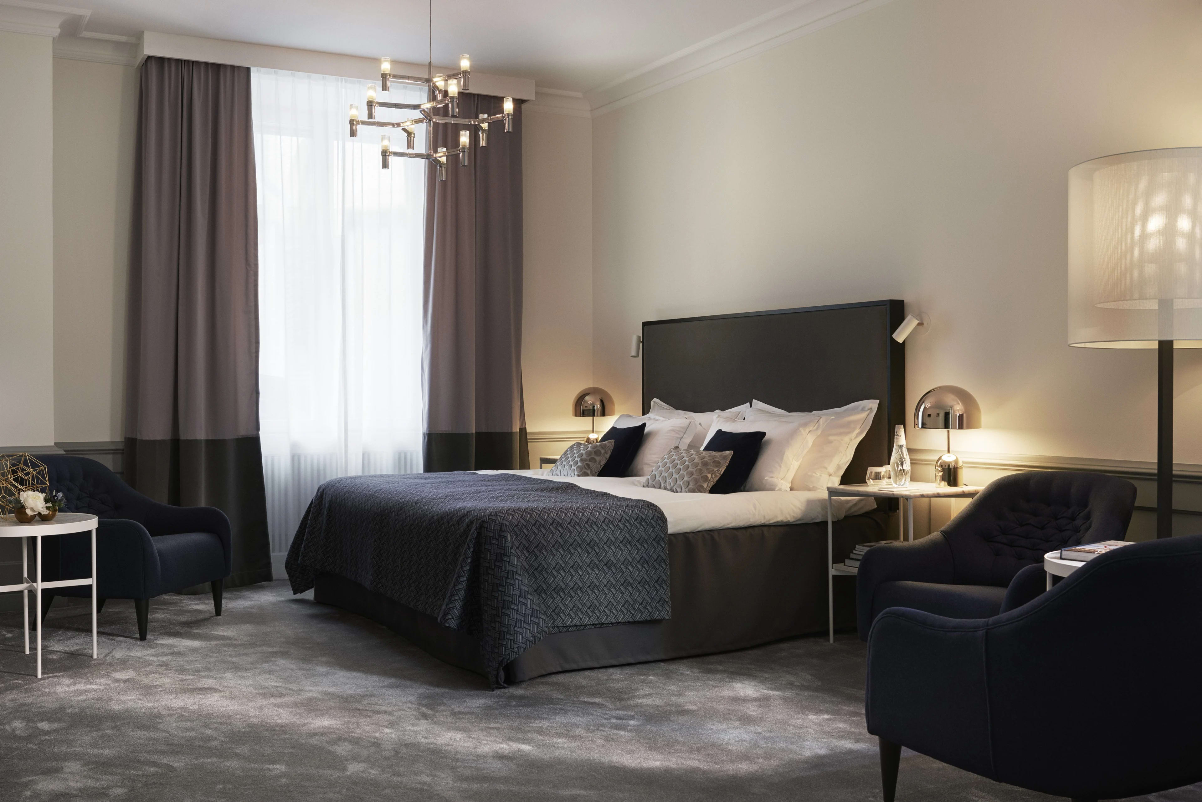 Modern hotel room with dark a blue bed and chair, grey carpet, modern chandelier, and white walls
