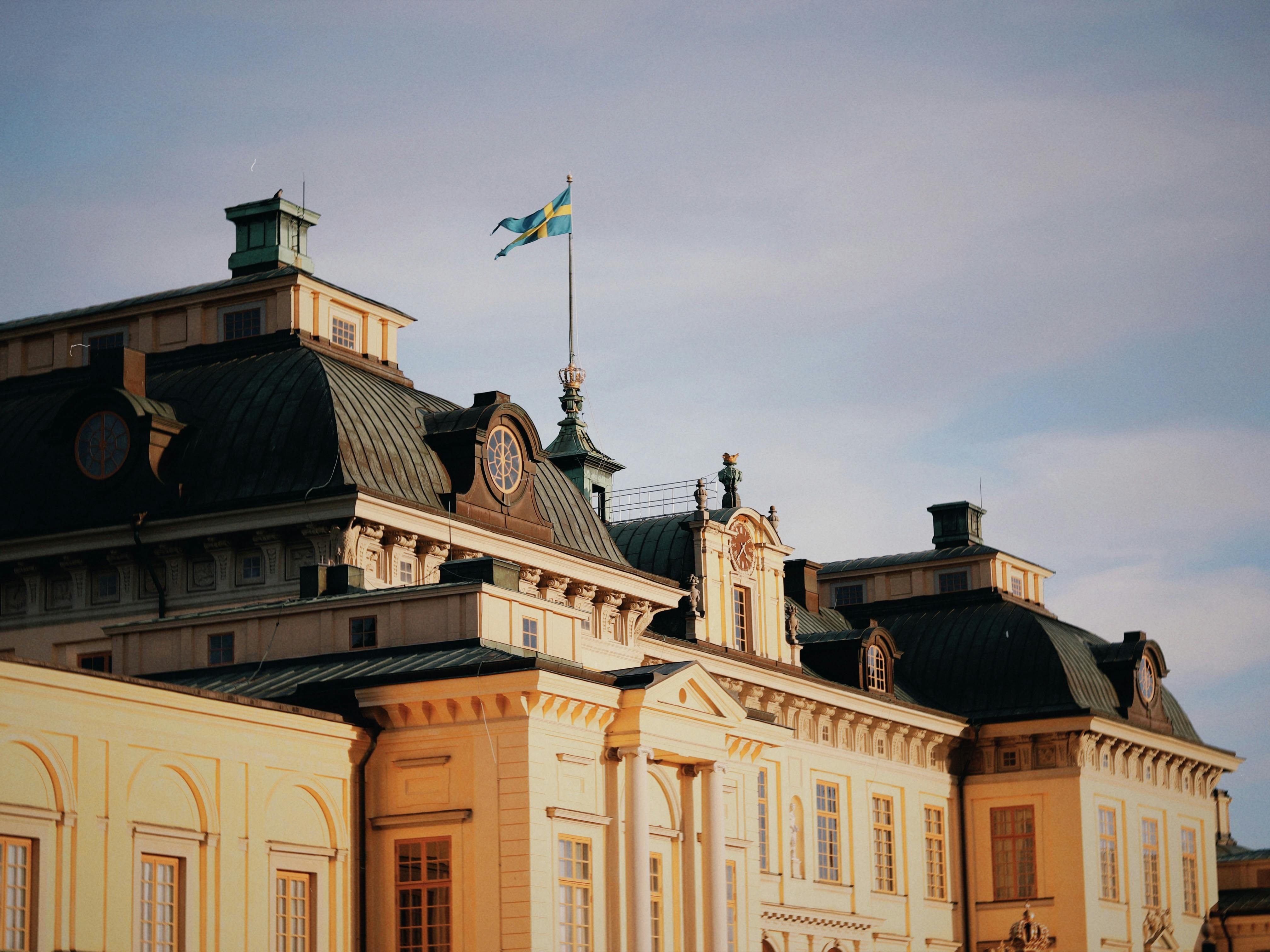a yellow castle facade with sloped grey roof and swedish flag during day