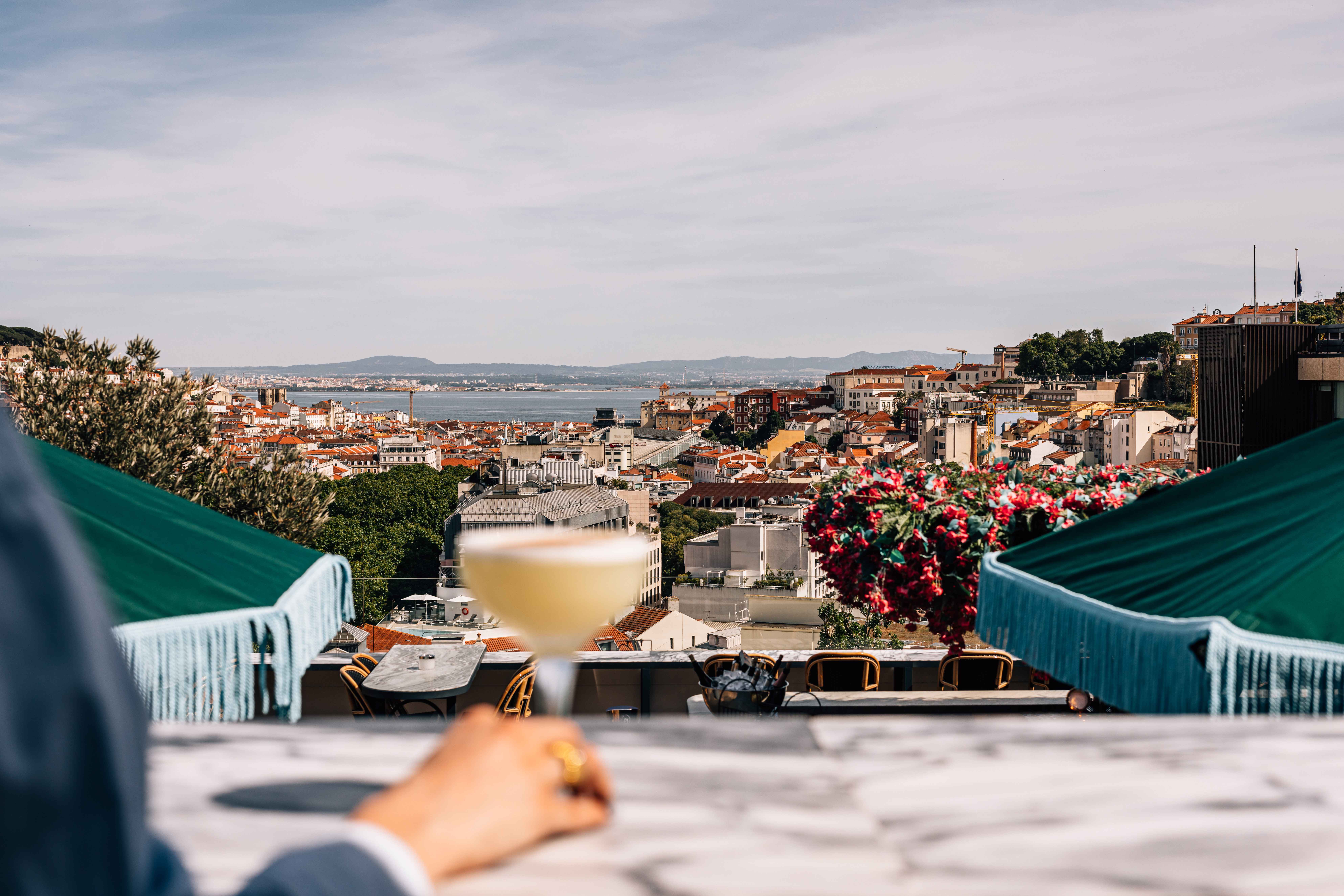 A beautiful view of the city of Lisbon with the ocean in the distance from a rooftop, showing a hand holding a cocktail.