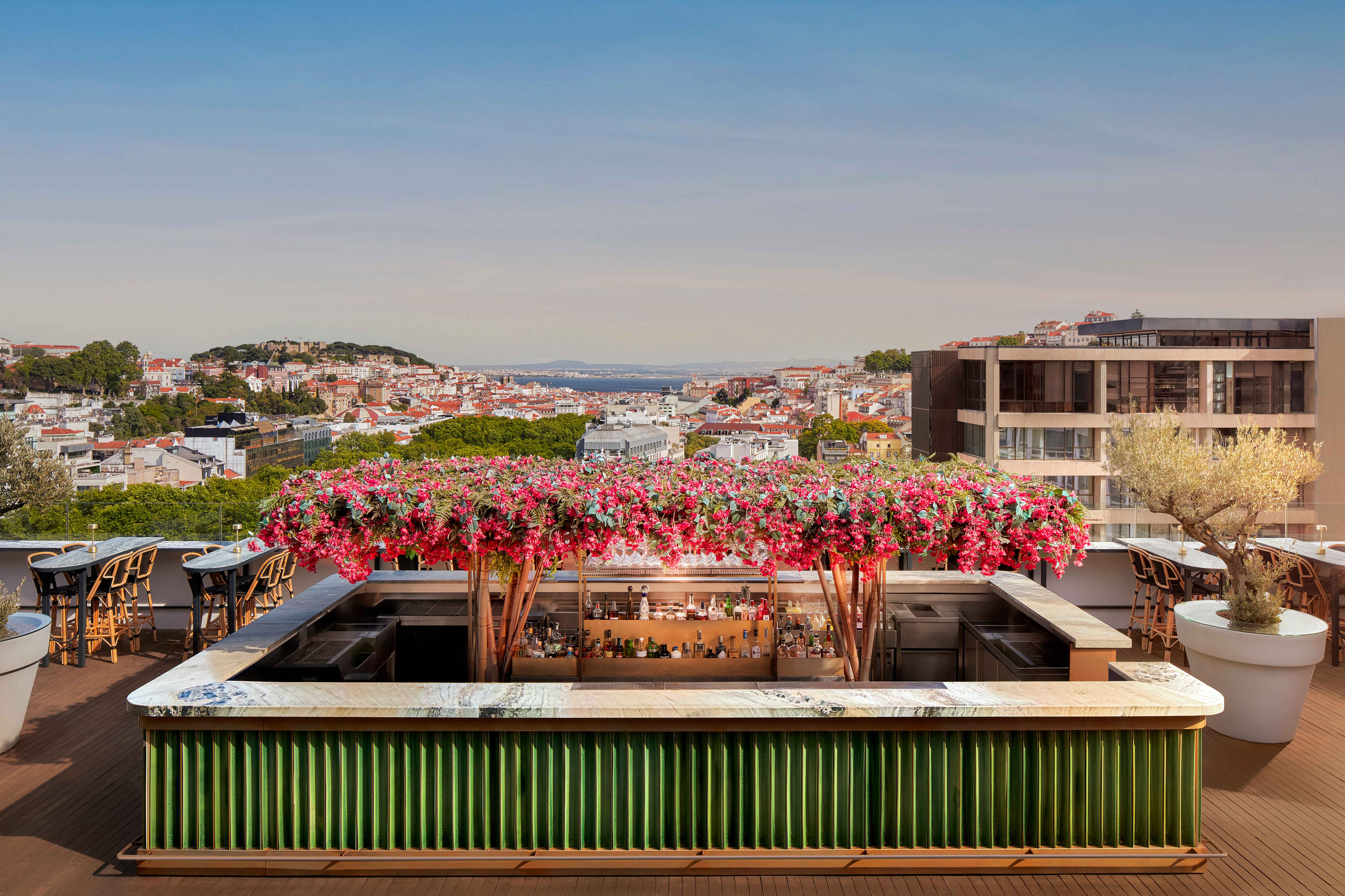 A rooftop with a striking, rectangular bar area in green and white with pink flowers overhead and city views.