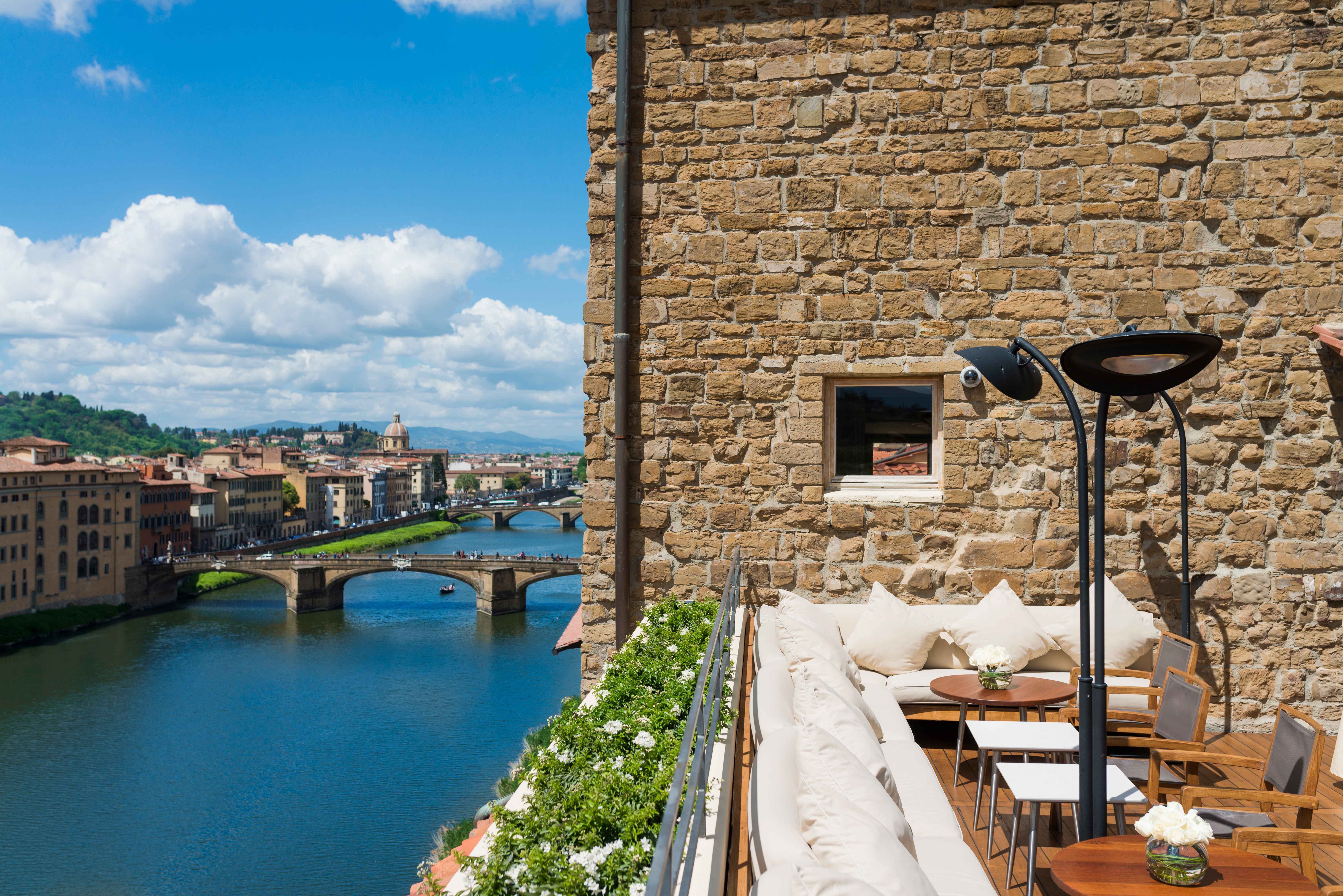 A view of a rooftop terrace with cream lounge seating around the edge against a stone wall and overlooking a river and bridge