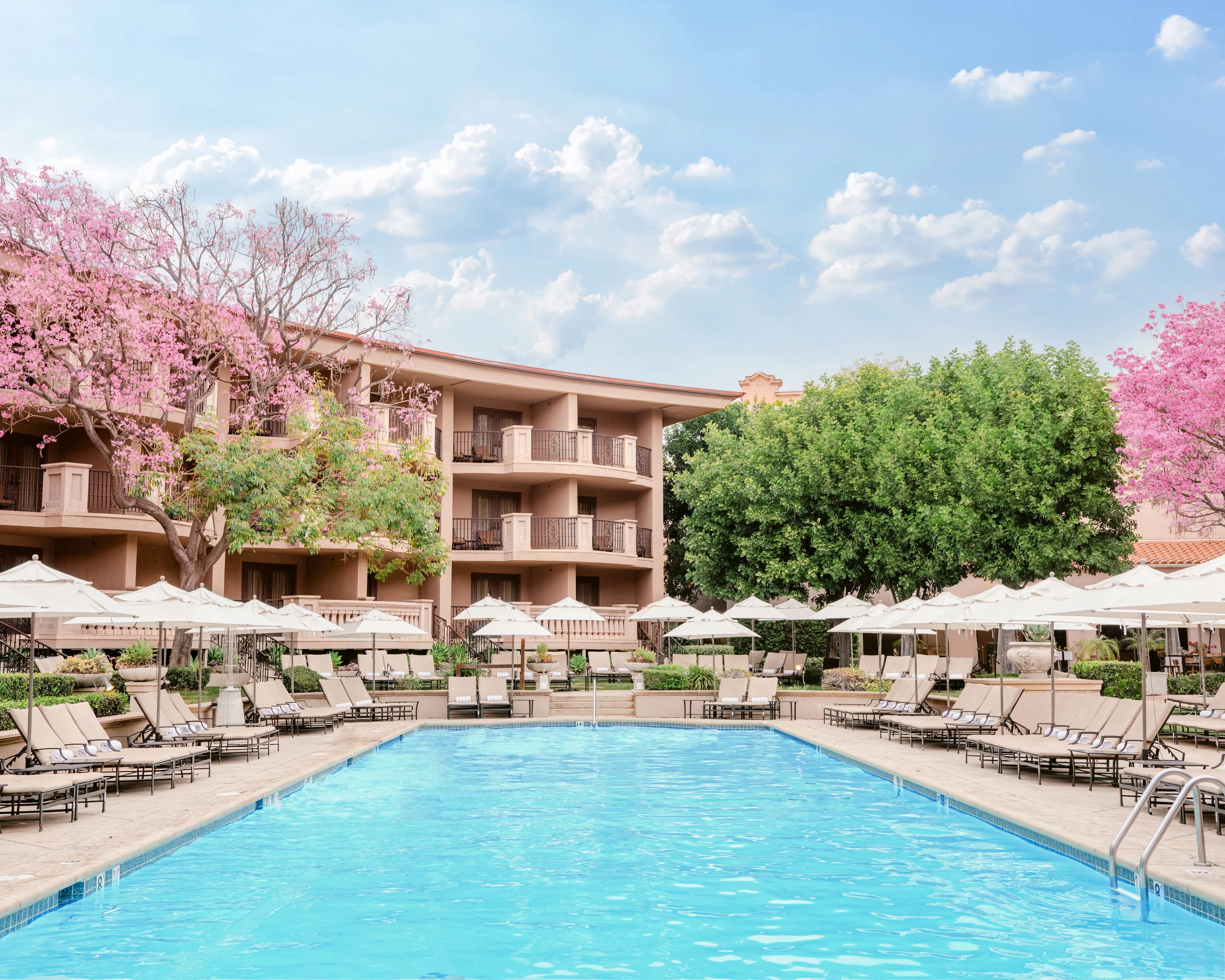a hotel pool with turquoise water, tan loungers, green trees, pink flowers, and brick hotel balconies during day