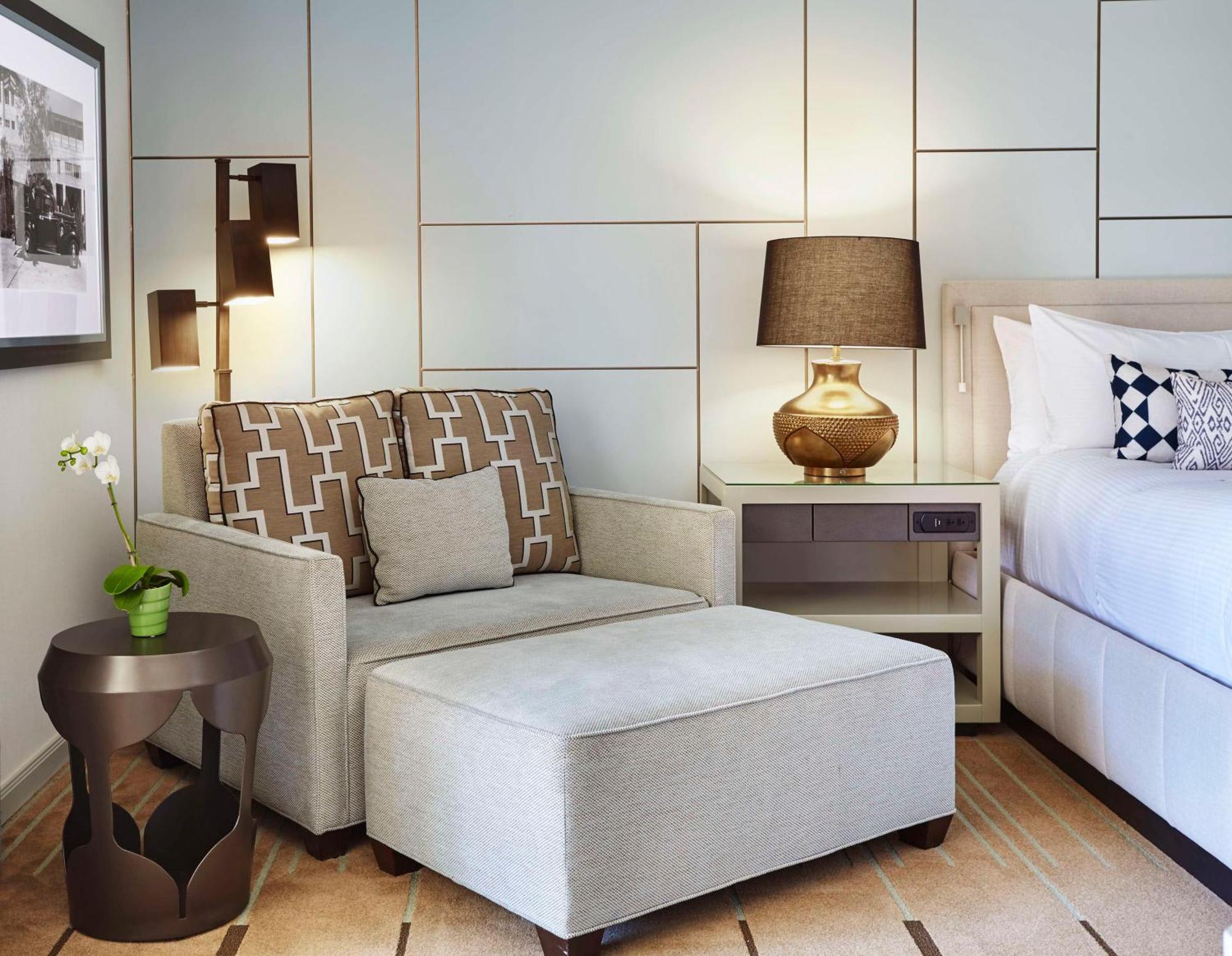 a white side chair and ottoman with brown pillows, bronze metal table and next to a hotel bed