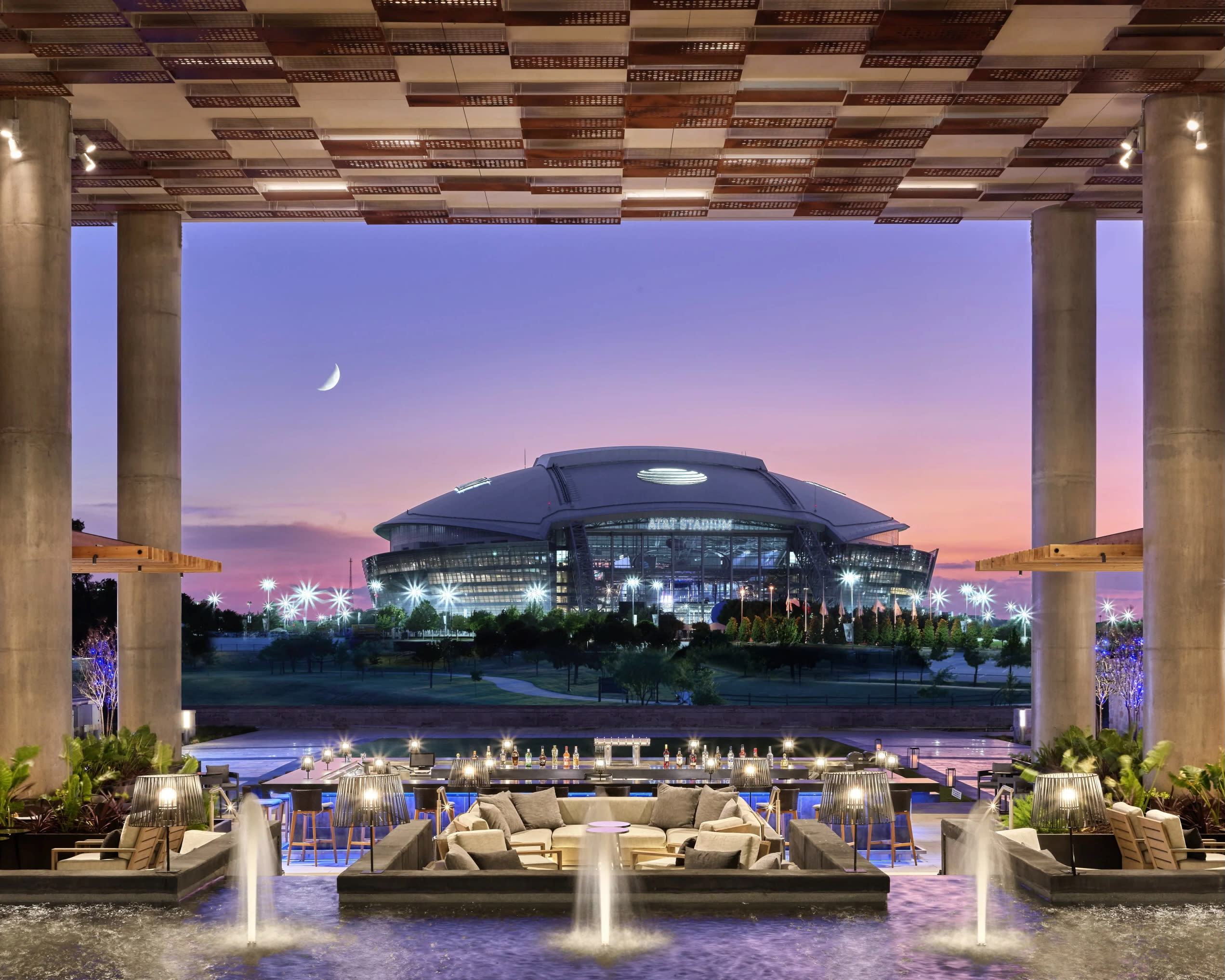 An outdoor hotel lounge with tall ceilings overlooking a football stadium arena at dusk
