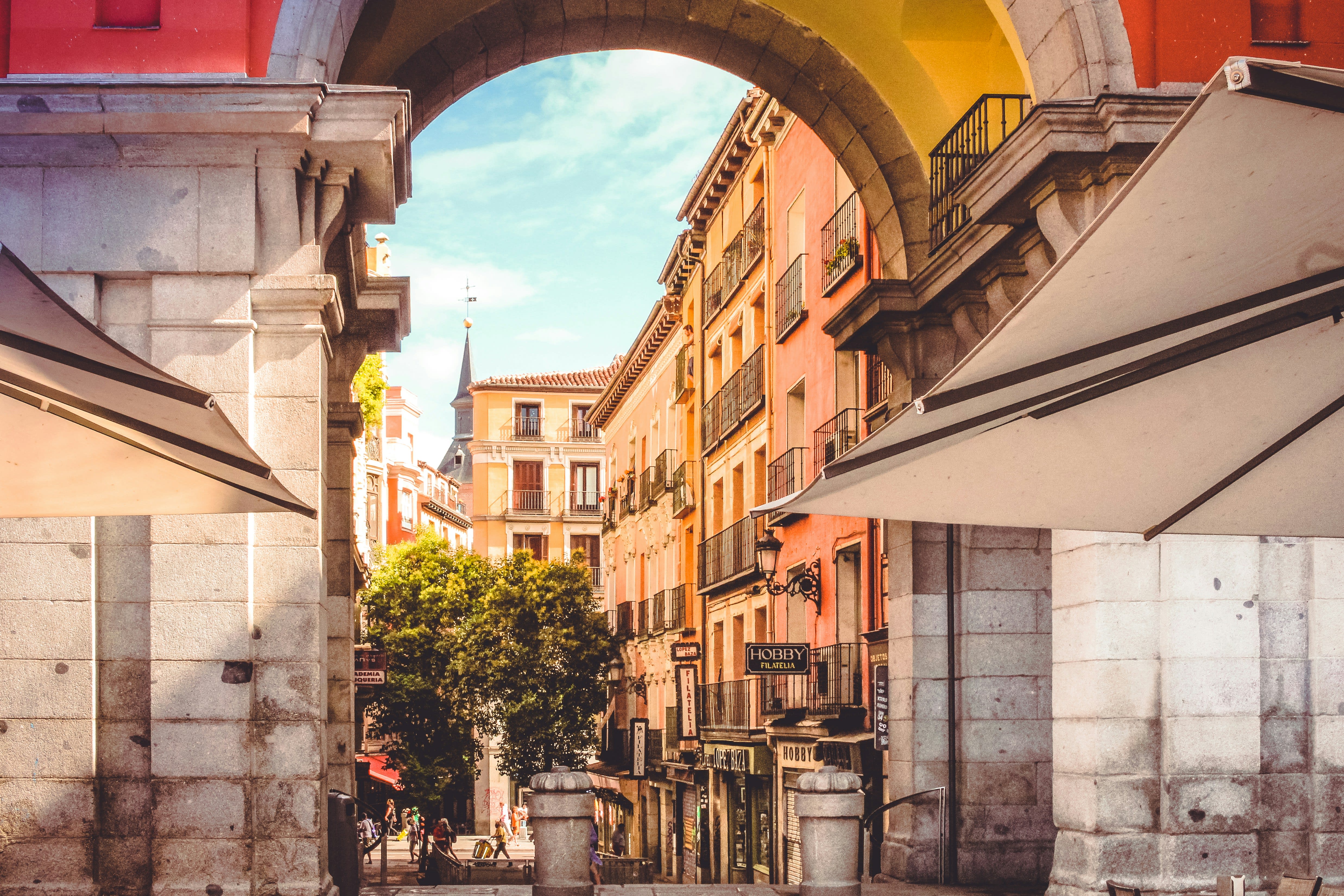 Archway view into a colorful street with balconies, shops, and a church spire in Madrid, Spain.