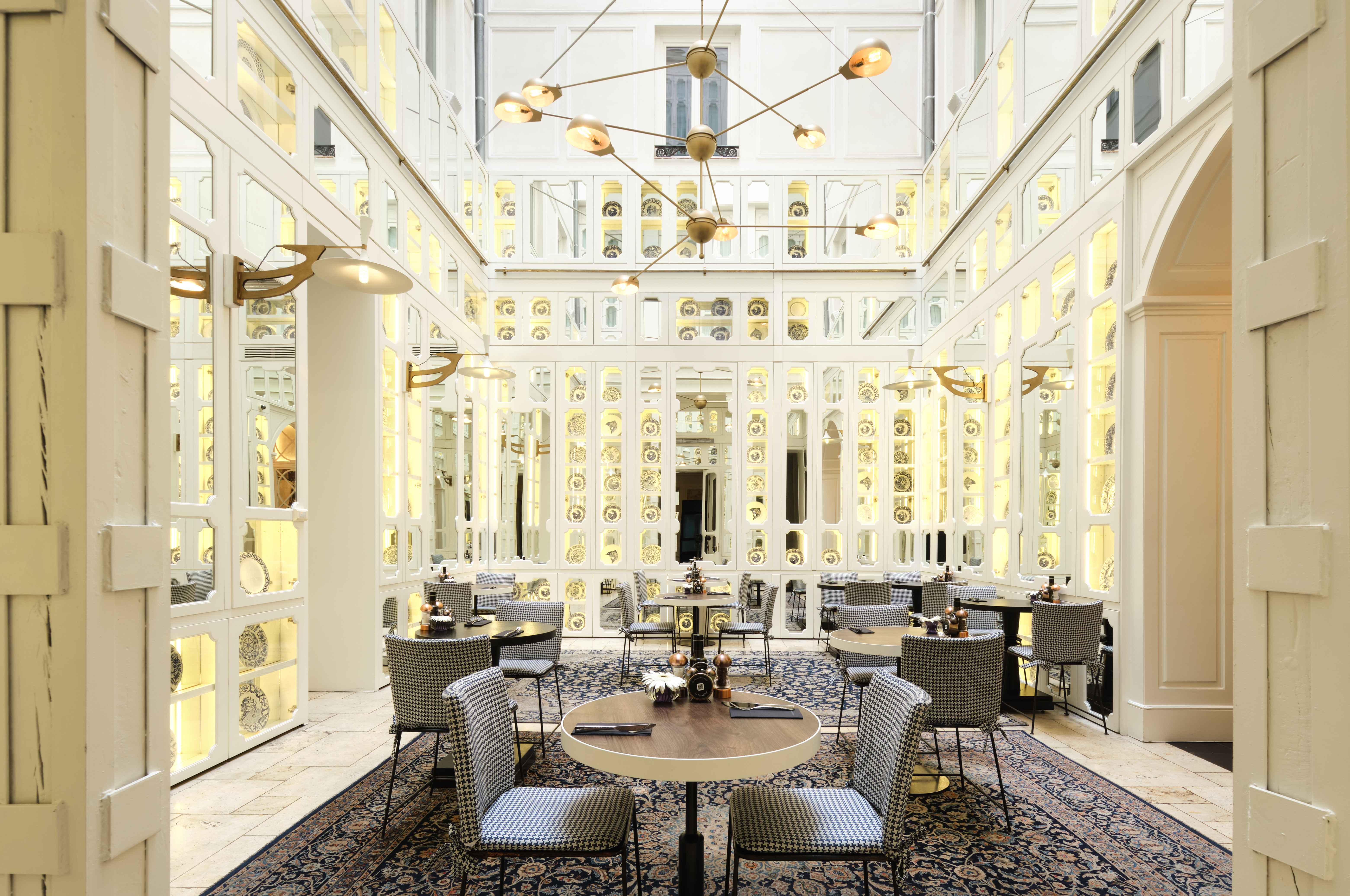 Bright restaurant with patterned chairs, round tables, and mirrored walls showcasing decorative plates under warm modern lighting.