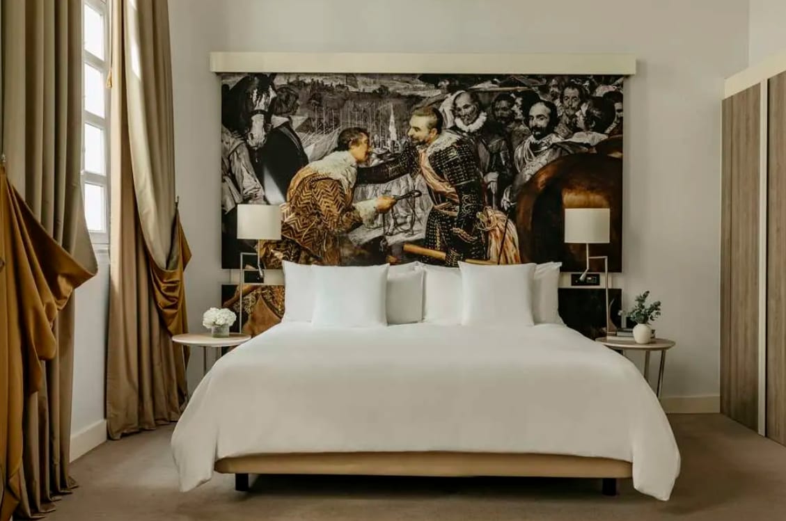 Elegant hotel room with a large bed, golden drapes, and a dramatic headboard mural depicting a historic painting scene.