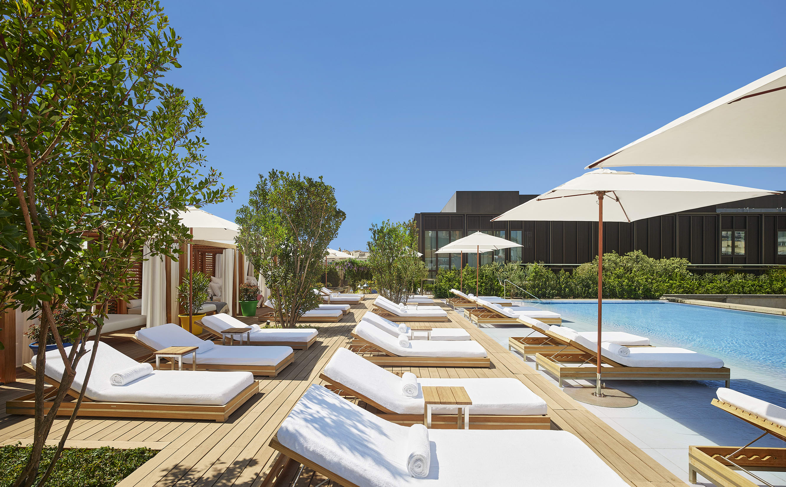 Outdoor pool deck with white sun loungers, umbrellas, and cabanas surrounded by greenery under a clear blue sky.