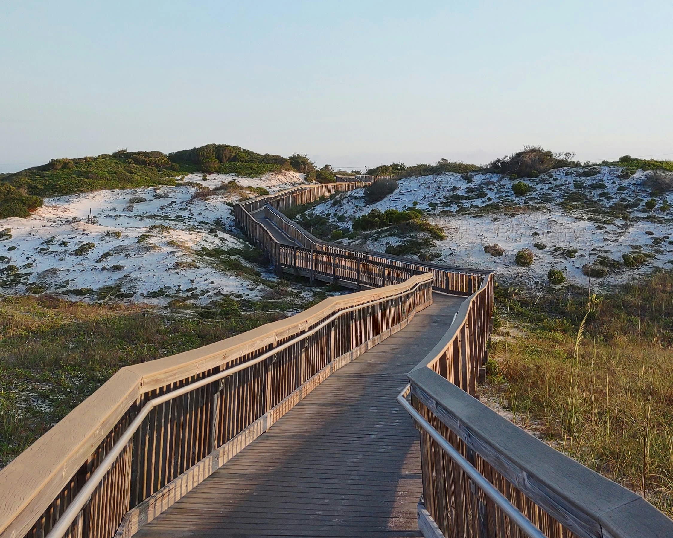 a wood walkway winding over beach grass toward a sand dune during day