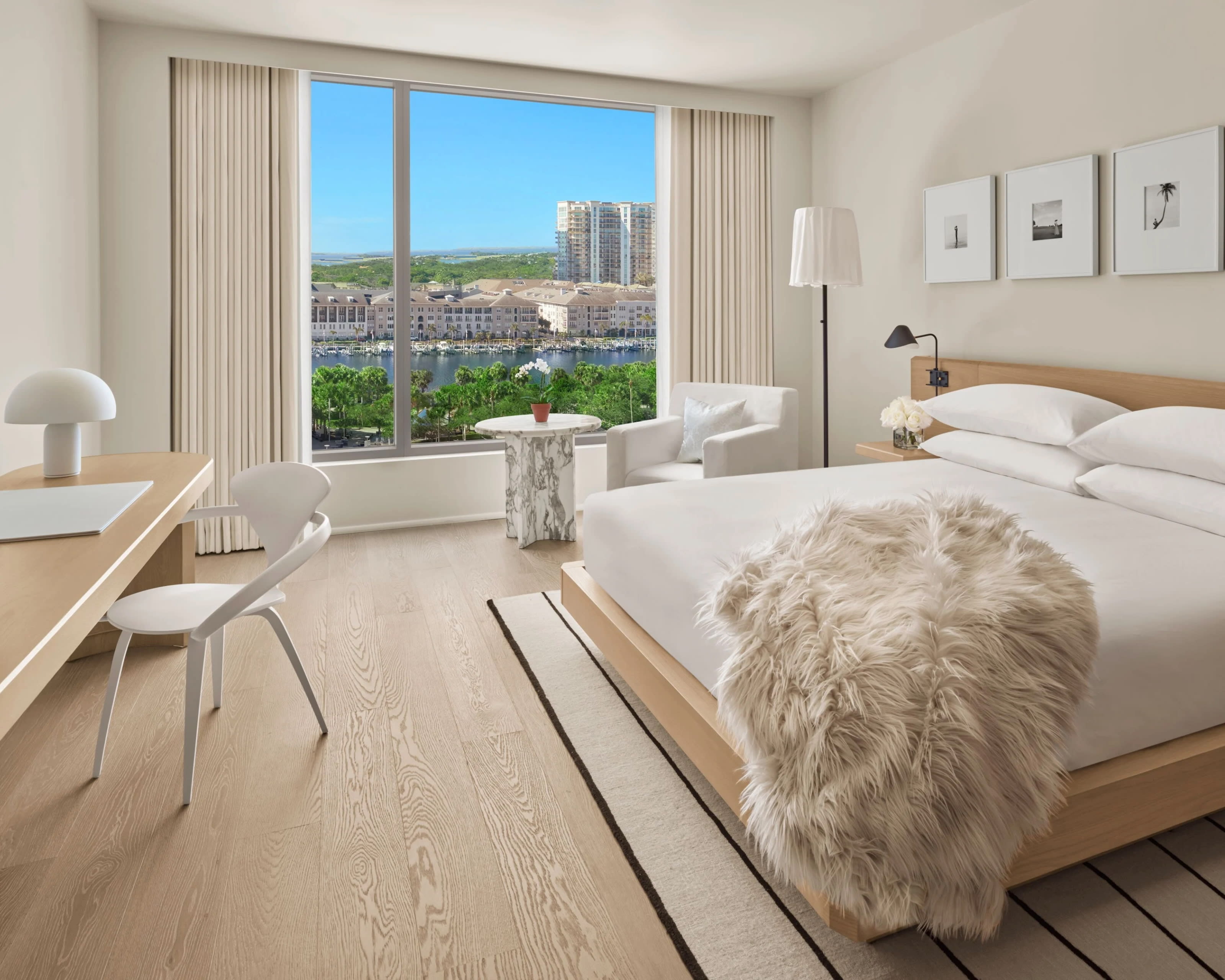 a modern hotel room with light wood floors and furniture, white linens and chair, and beige fur throw on the bed