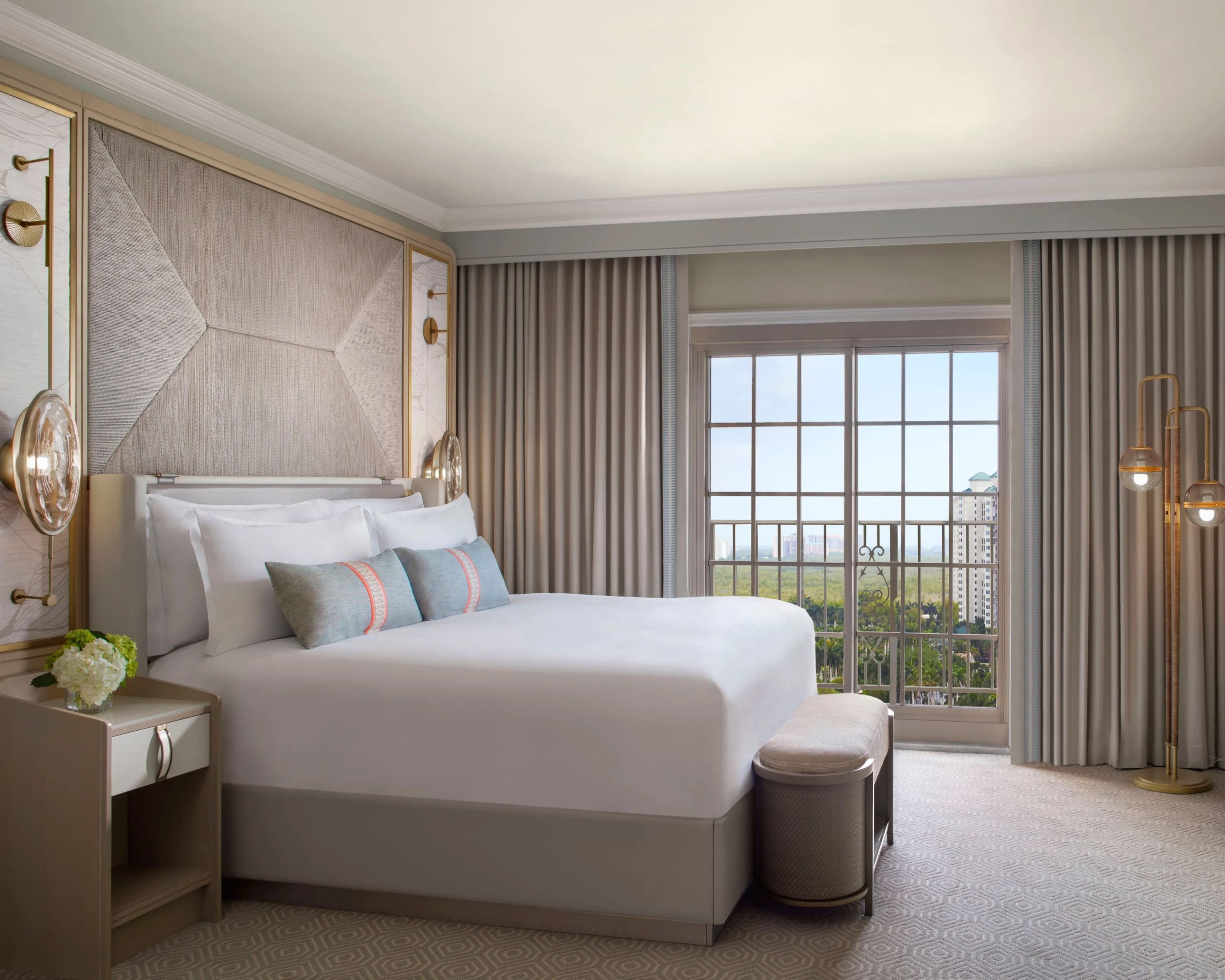 Beige hotel room with light blue and pink pillows on the bed, designed headboard and sliding glass doors to a balcony