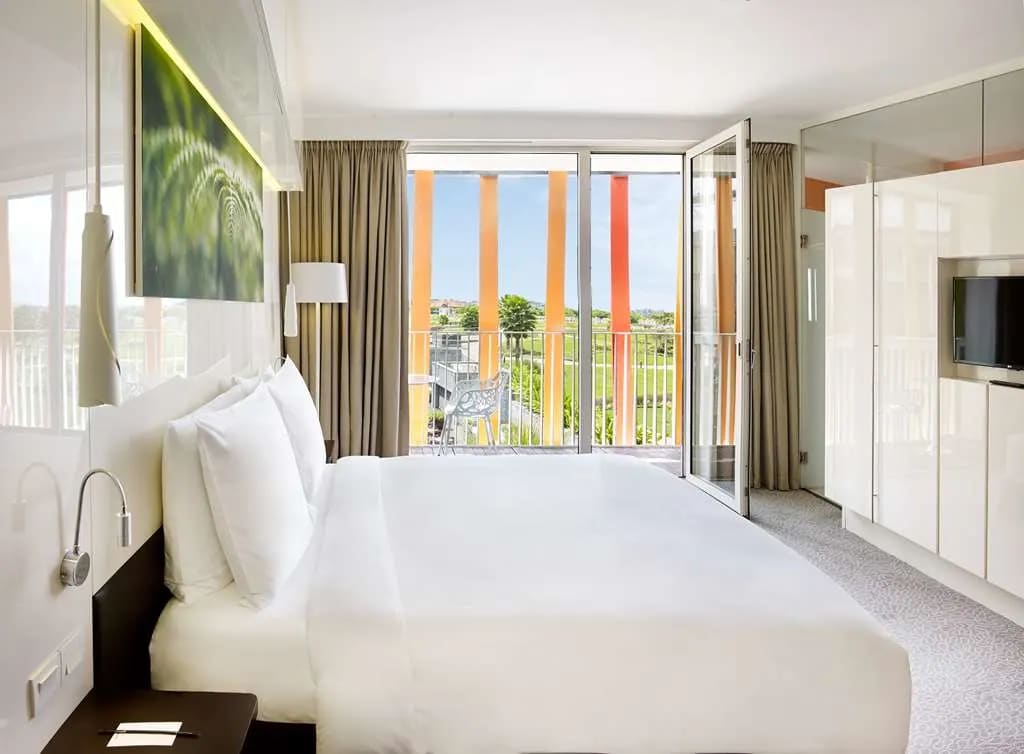 white hotel bed with green landscape photo above bed and windows looking outside with bright orange modern metal siding