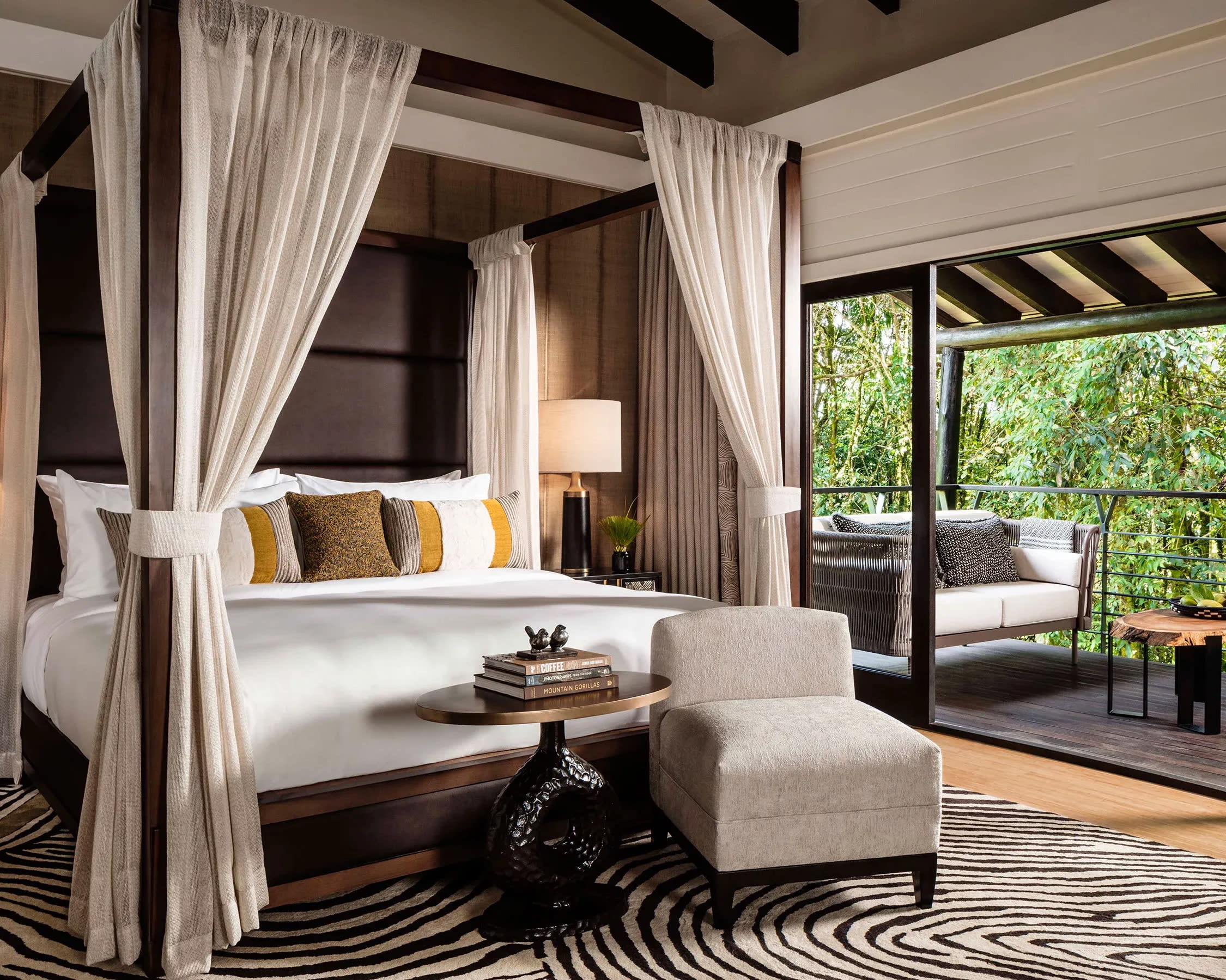 A white hotel bed with dark wood four poster bed, zebra pattern rug, and balcony with green trees beyond