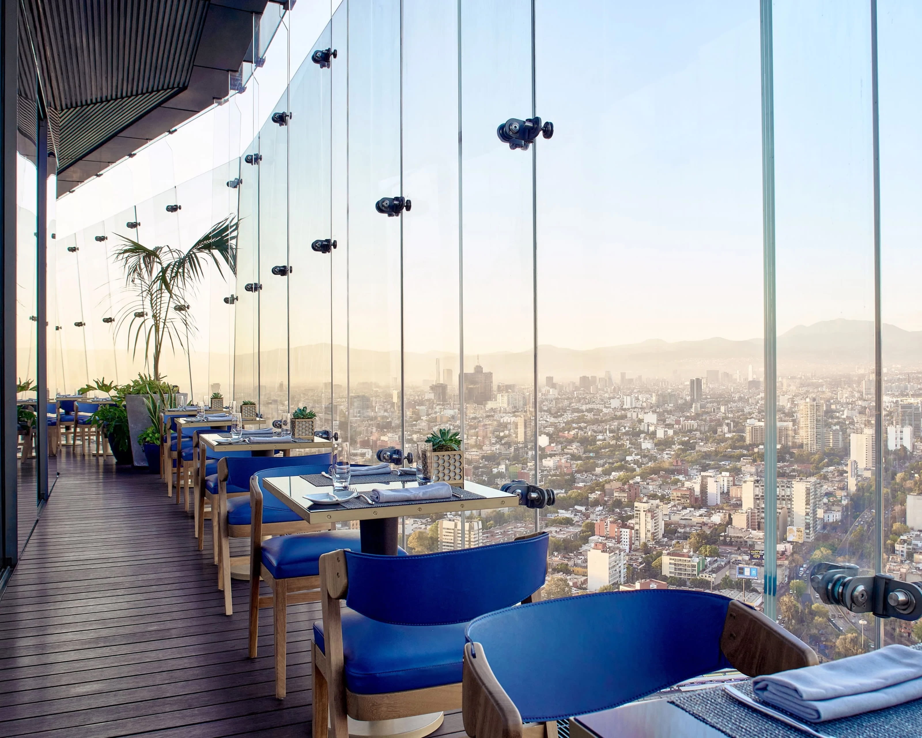 blue chairs and wood tables on an outdoor patio with glass windows overlooking a cityscape