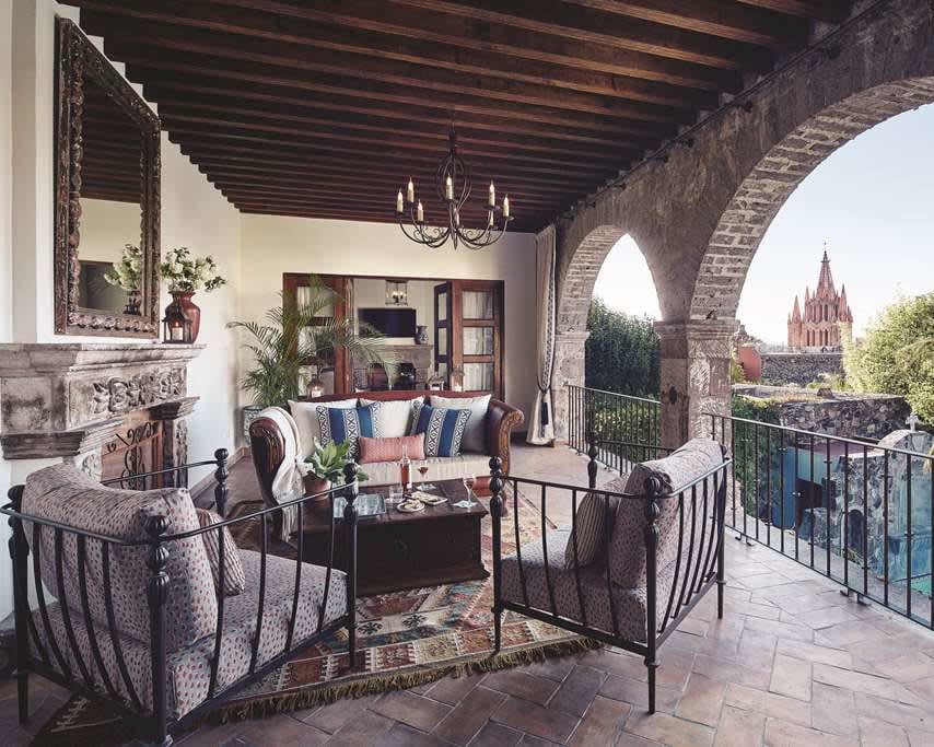 a colonial style balcony with outdoor furniture, chandelier, wood beam ceiling, and arches with railings and a tower in the background