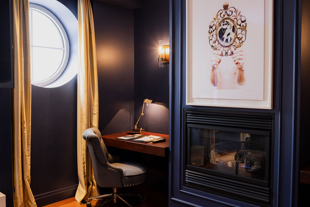 Cozy corner with a writing desk, round window, and a fireplace set into a navy wall topped with modern artwork.