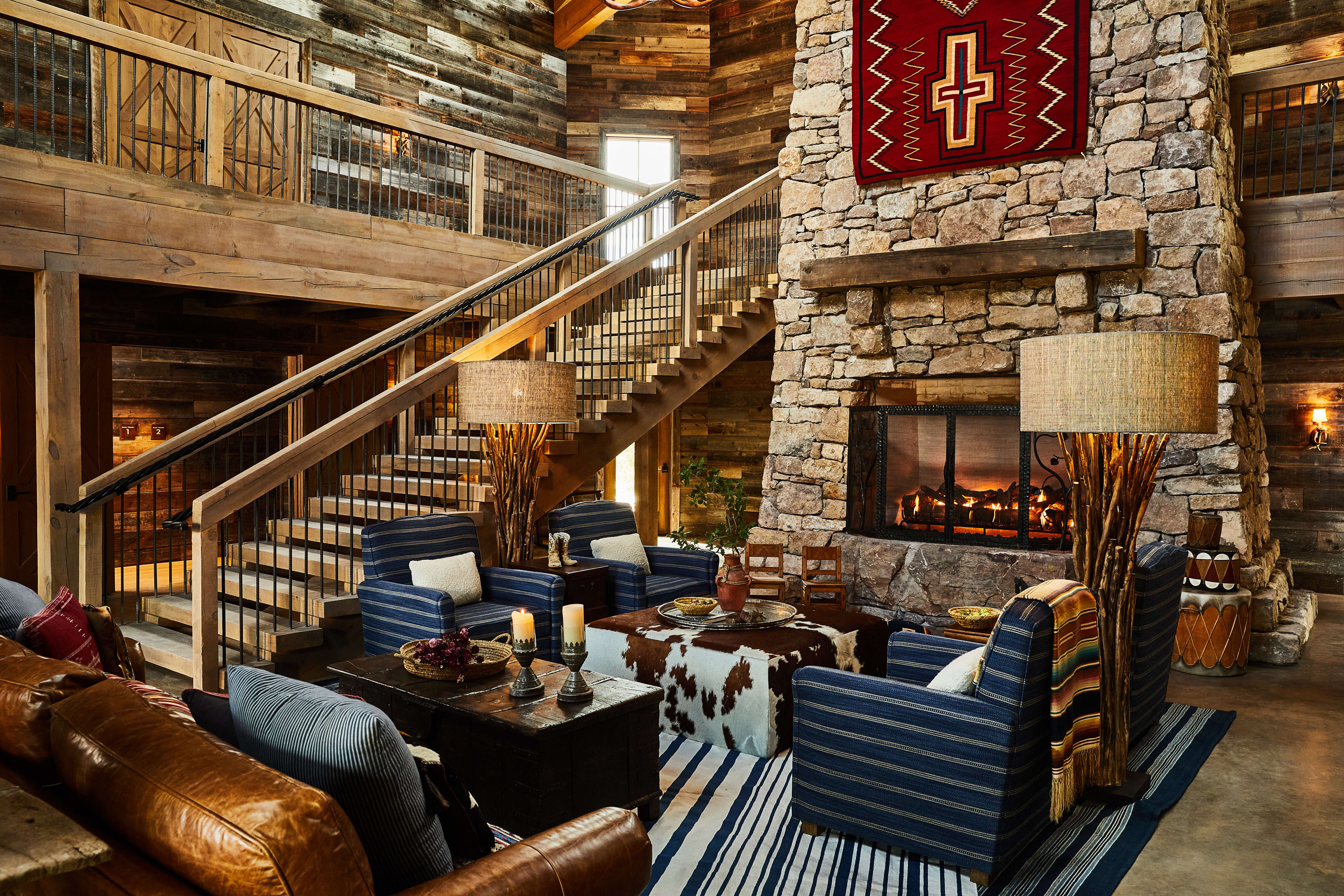 Rustic lodge living room with a stone fireplace, leather and striped armchairs, wooden beams, and a staircase leading to a loft.