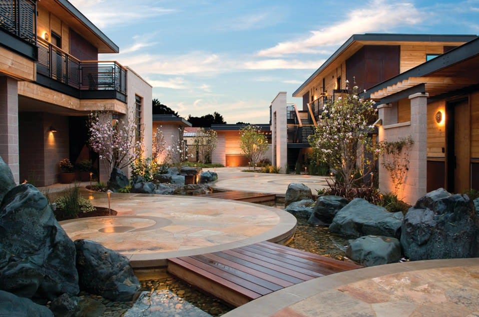 Modern courtyard with stone paths, water features, blossoming trees, and contemporary buildings at sunset.