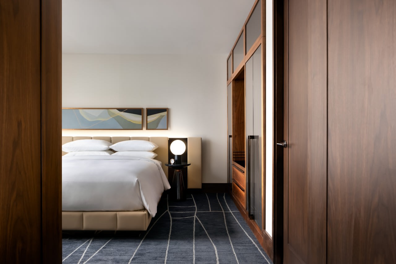 Modern hotel bedroom with a sleek design, featuring a white bed with crisp linens, abstract artwork above the headboard, a round bedside lamp, and wooden paneling with built-in storage.