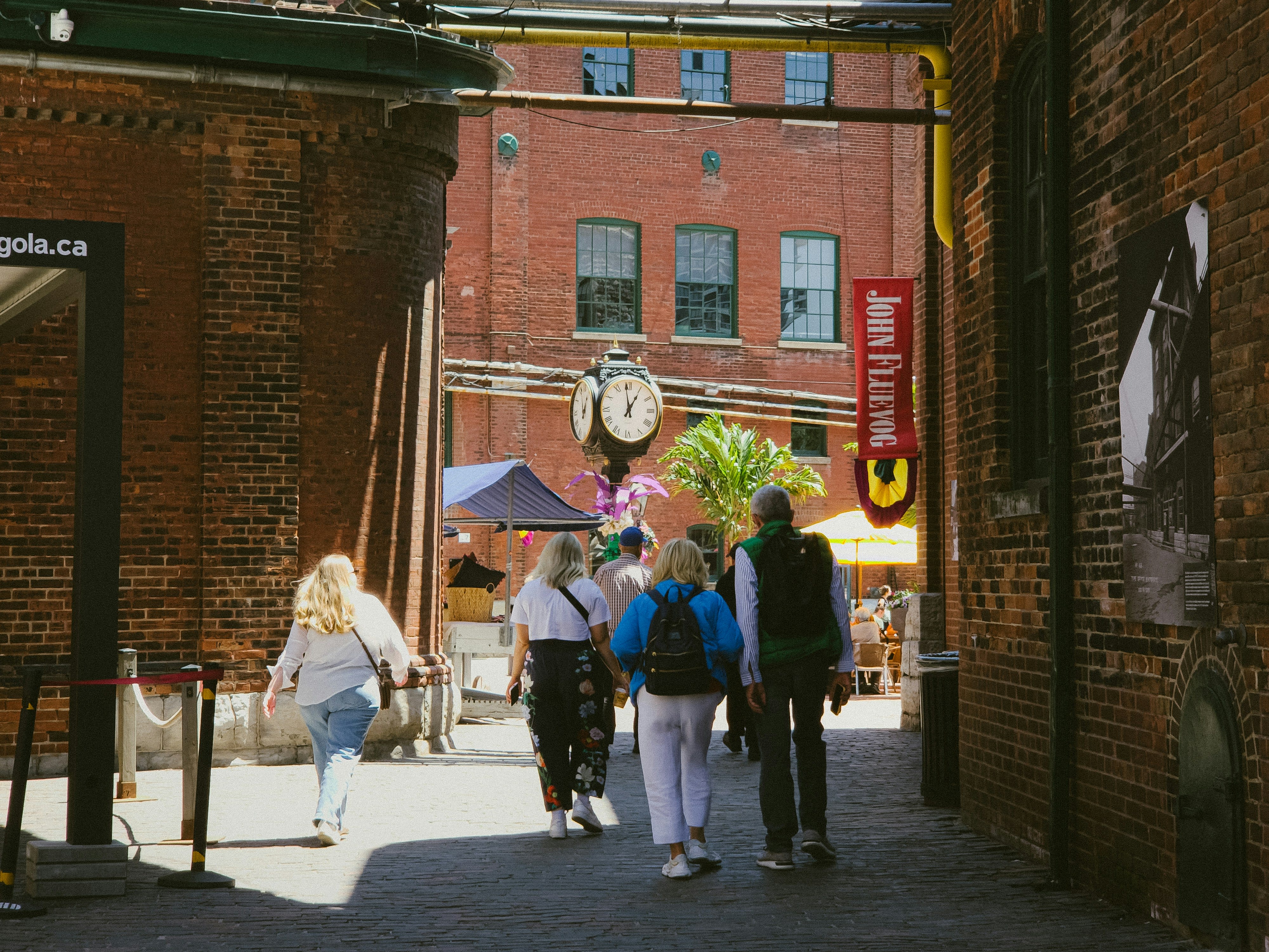 People walking through Toronto’s Distillery District, surrounded by red-brick buildings, a vintage street clock, and colorful outdoor market stalls.