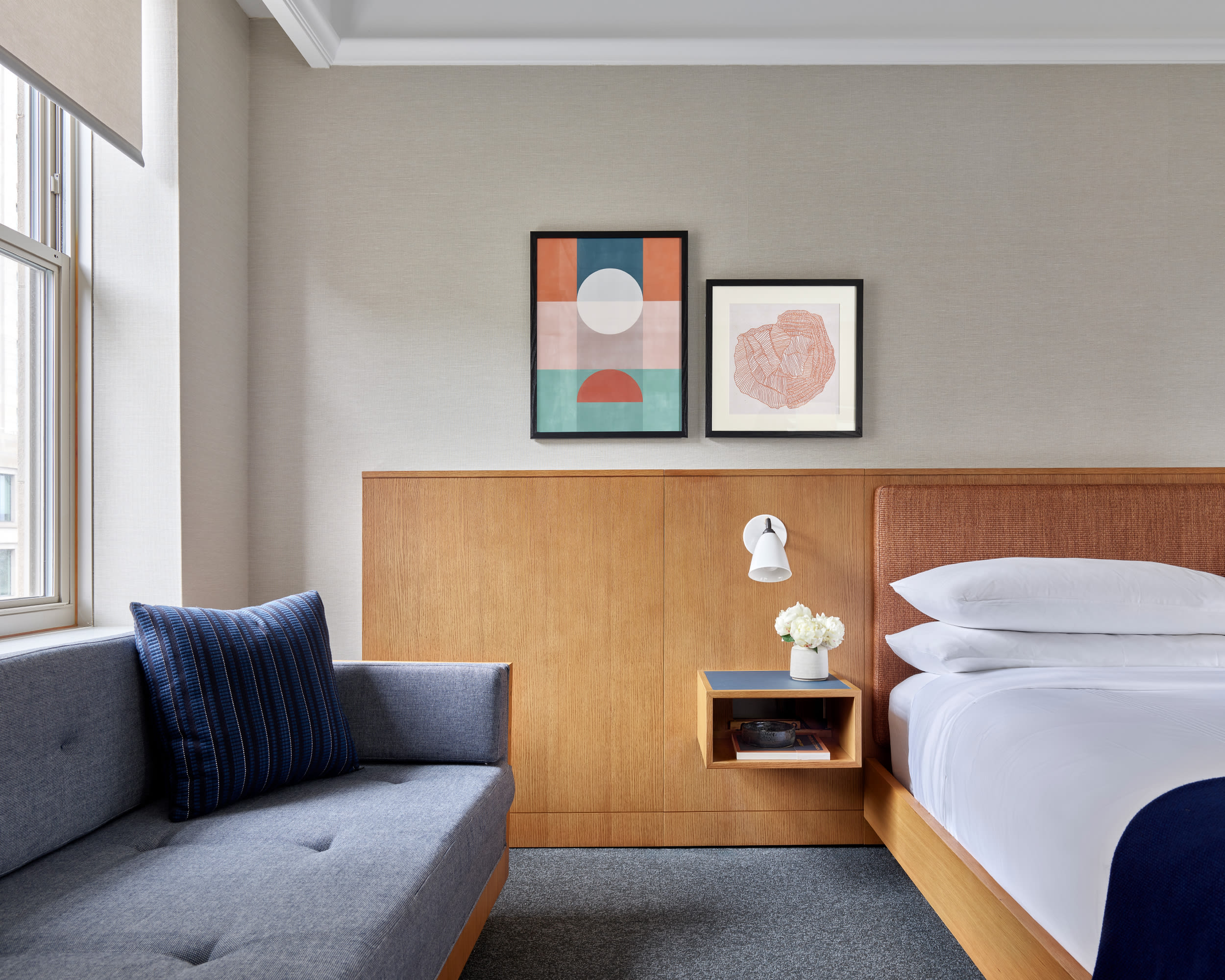 Modern hotel room with a minimalist design featuring a wooden bed frame with crisp white bedding, a floating bedside table with flowers, a gray sofa with a blue cushion, and two framed artworks on the wall above the headboard.