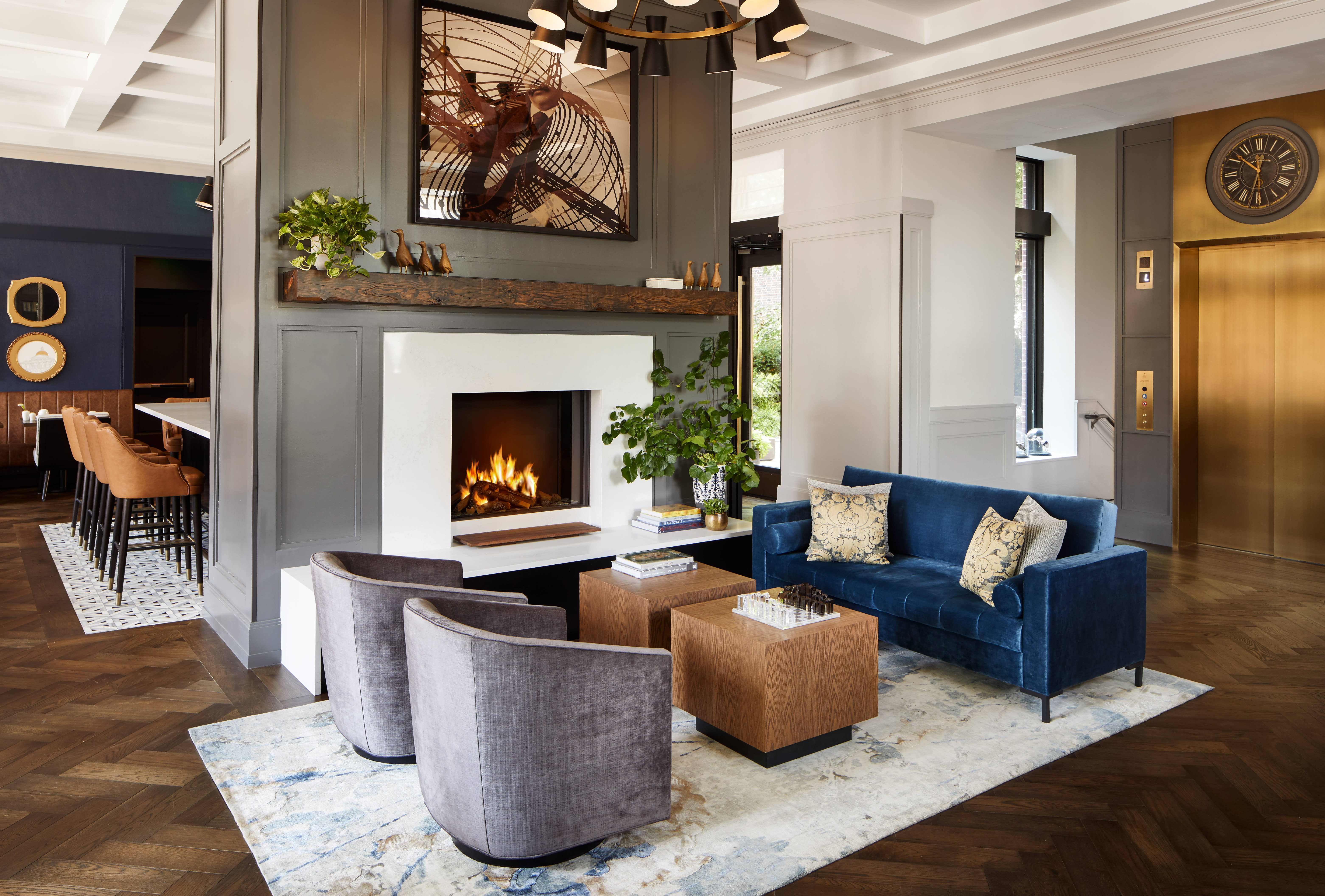 Modern hotel lobby featuring a sleek fireplace with a white surround, flanked by plush seating including a deep blue velvet sofa and two gray armchairs. Warm wood accents, a brass elevator door, and a stylish bar area with leather stools.