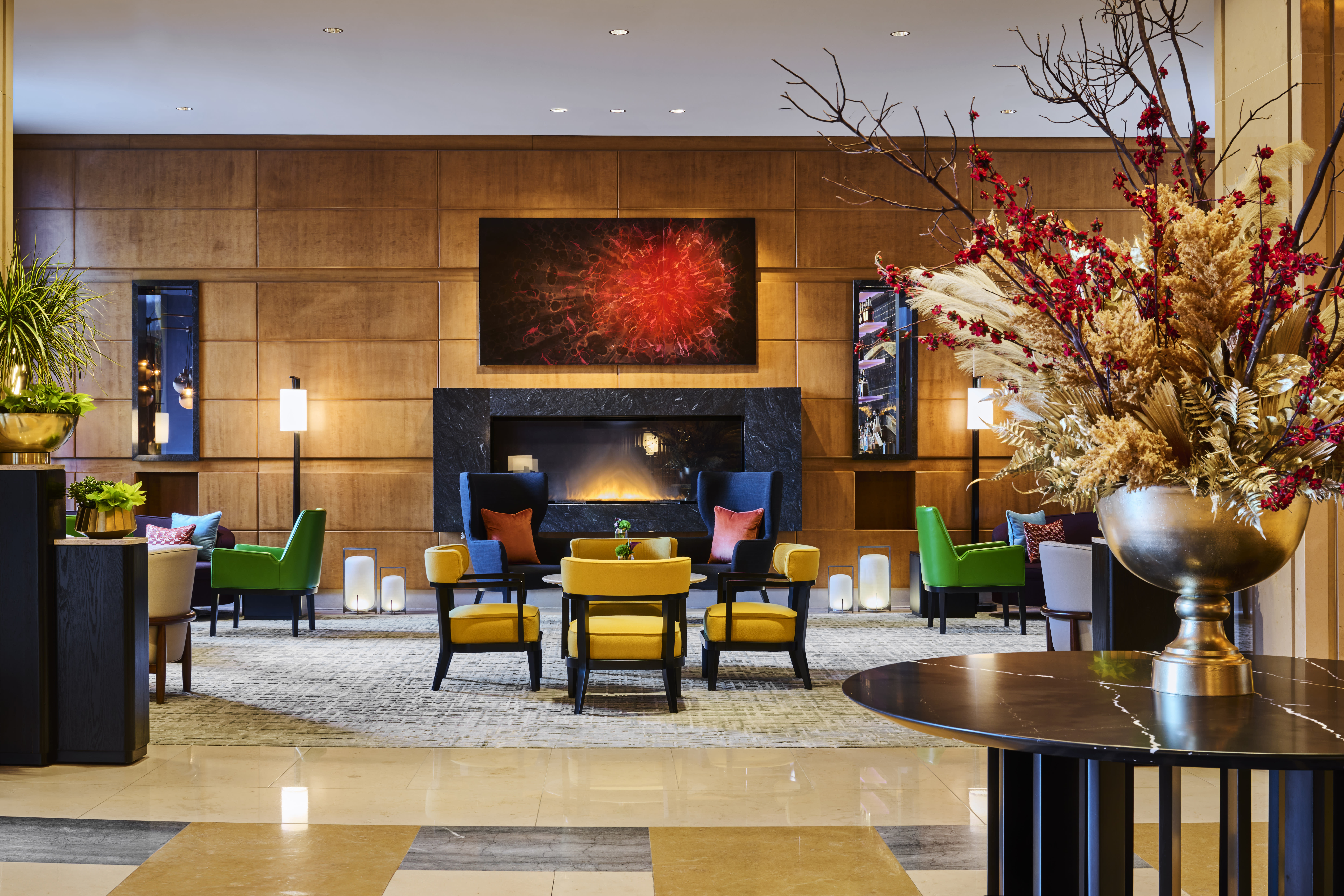 A vibrant hotel lobby with a modern fireplace set into a sleek black surround, framed by wood-paneled walls. Brightly colored chairs in yellow, green, and blue with accent pillows create a lively seating area