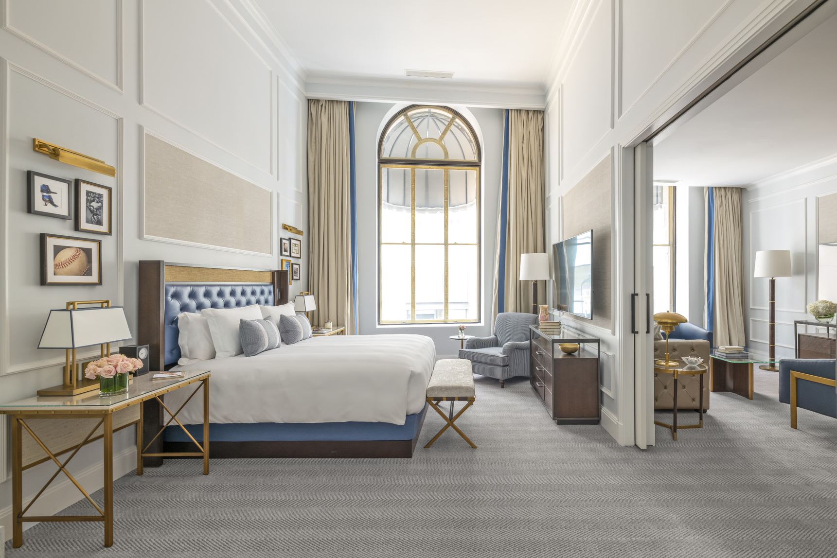 legant hotel suite with high ceilings and a grand arched window framed by floor-to-ceiling drapes. The bedroom features a king bed with a tufted navy headboard, flanked by gold-accented side tables and modern lamps.