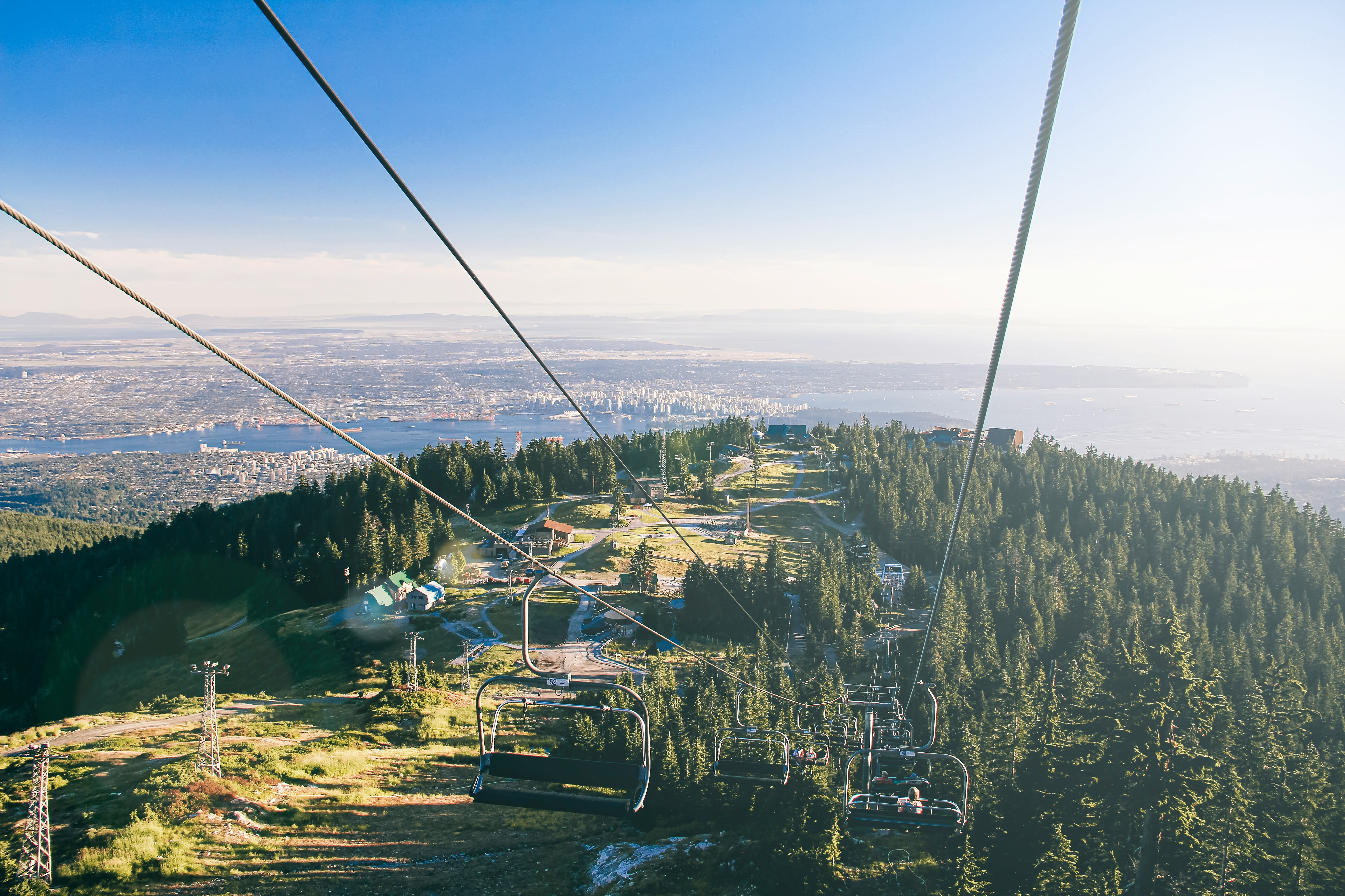 A scenic chairlift ride up Grouse Mountain overlooking dense evergreen forests, with sweeping views of Vancouver’s skyline, harbor, and distant coastline under a clear blue sky.
