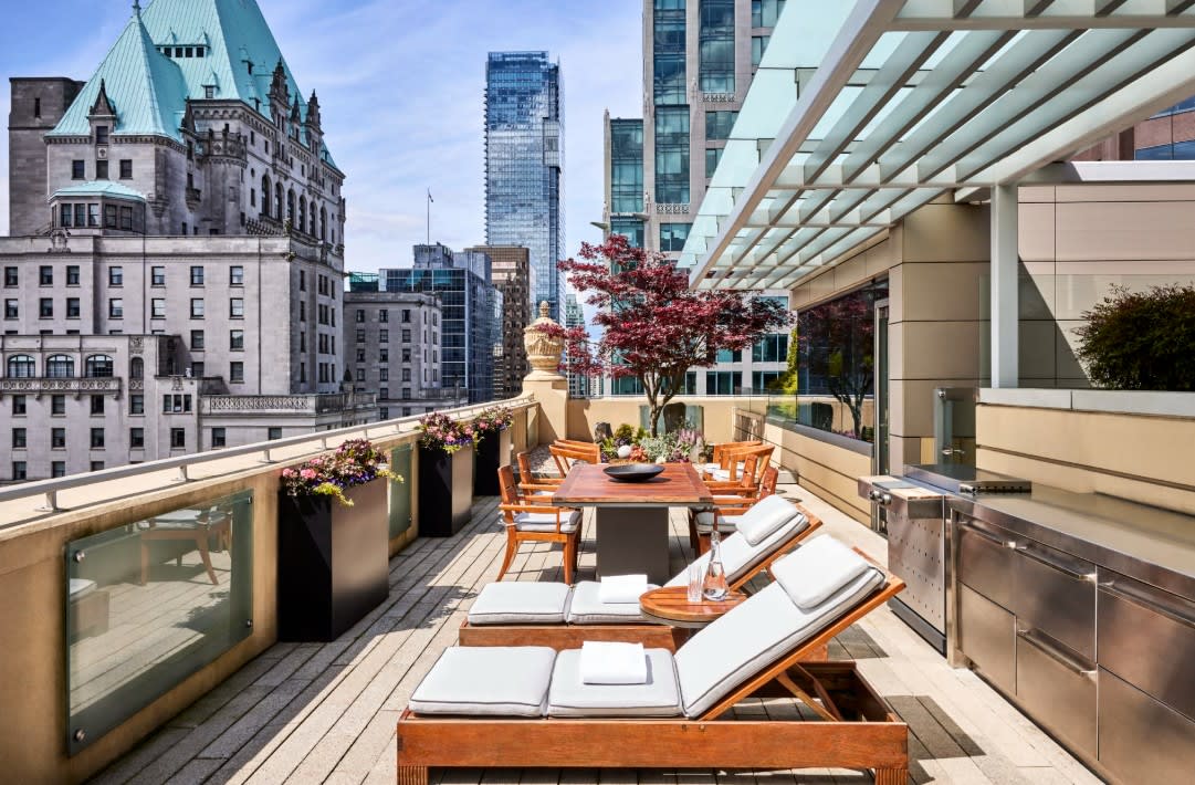 A stylish rooftop terrace with cushioned wooden loungers, a dining table surrounded by chairs, and a sleek outdoor kitchen, set against a backdrop of historic and modern high-rise buildings in downtown Vancouver.