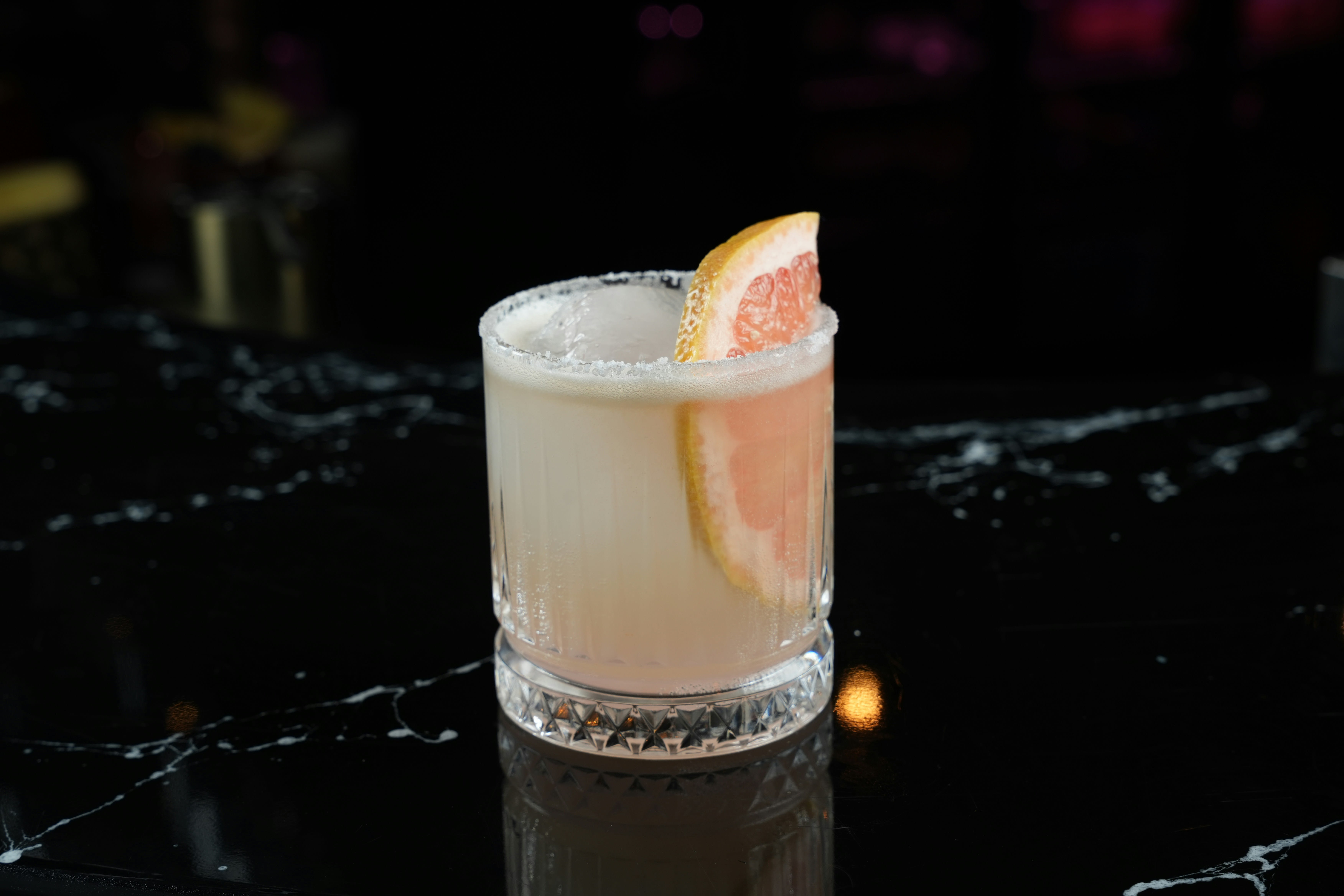 A refreshing cocktail served in a clear, textured glass with a salted rim, large ice cube, and a slice of grapefruit garnish