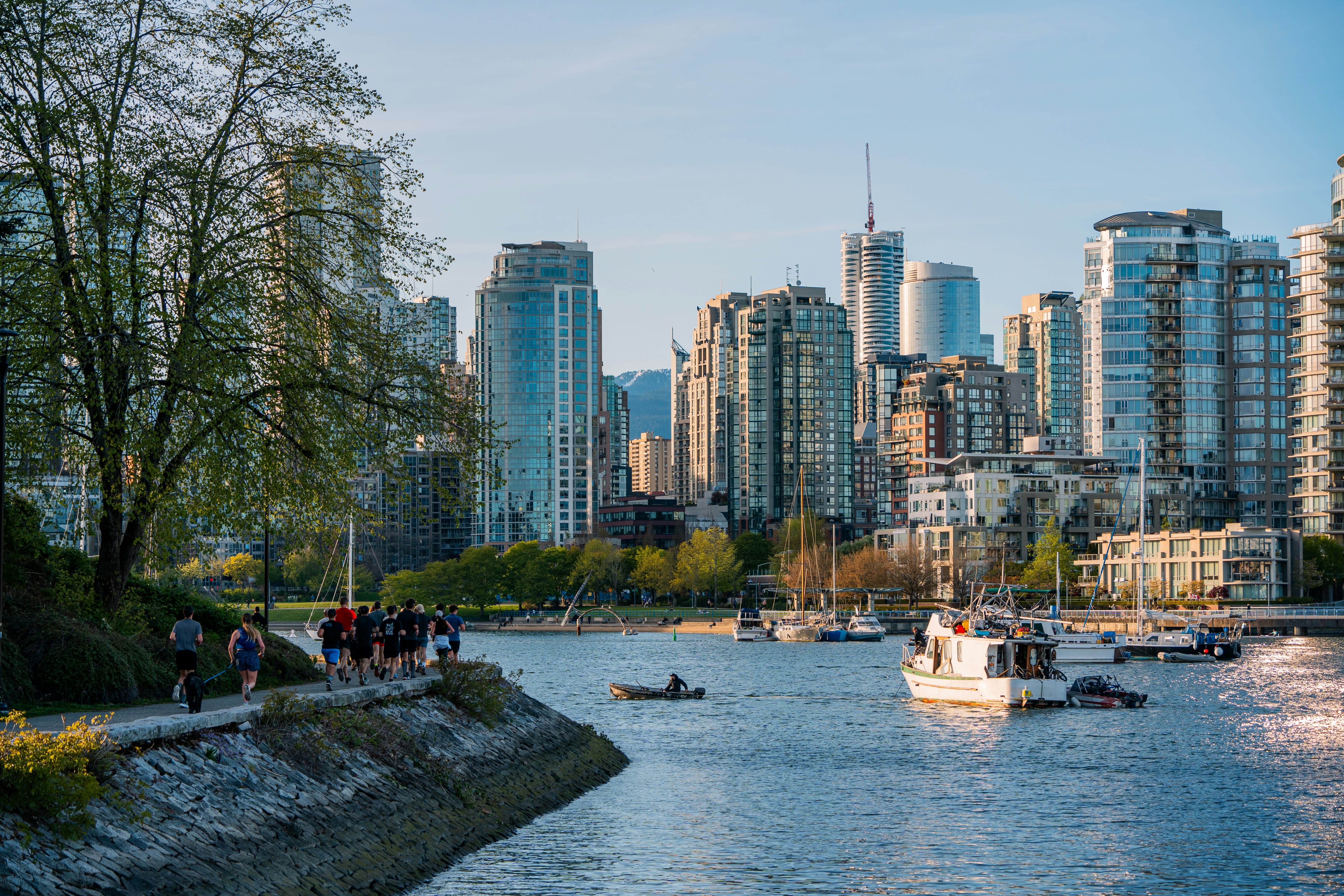 A lively Vancouver waterfront scene with people jogging along the seawall, boats anchored in the harbor, and modern high-rise buildings towering in the background under a clear blue sky.