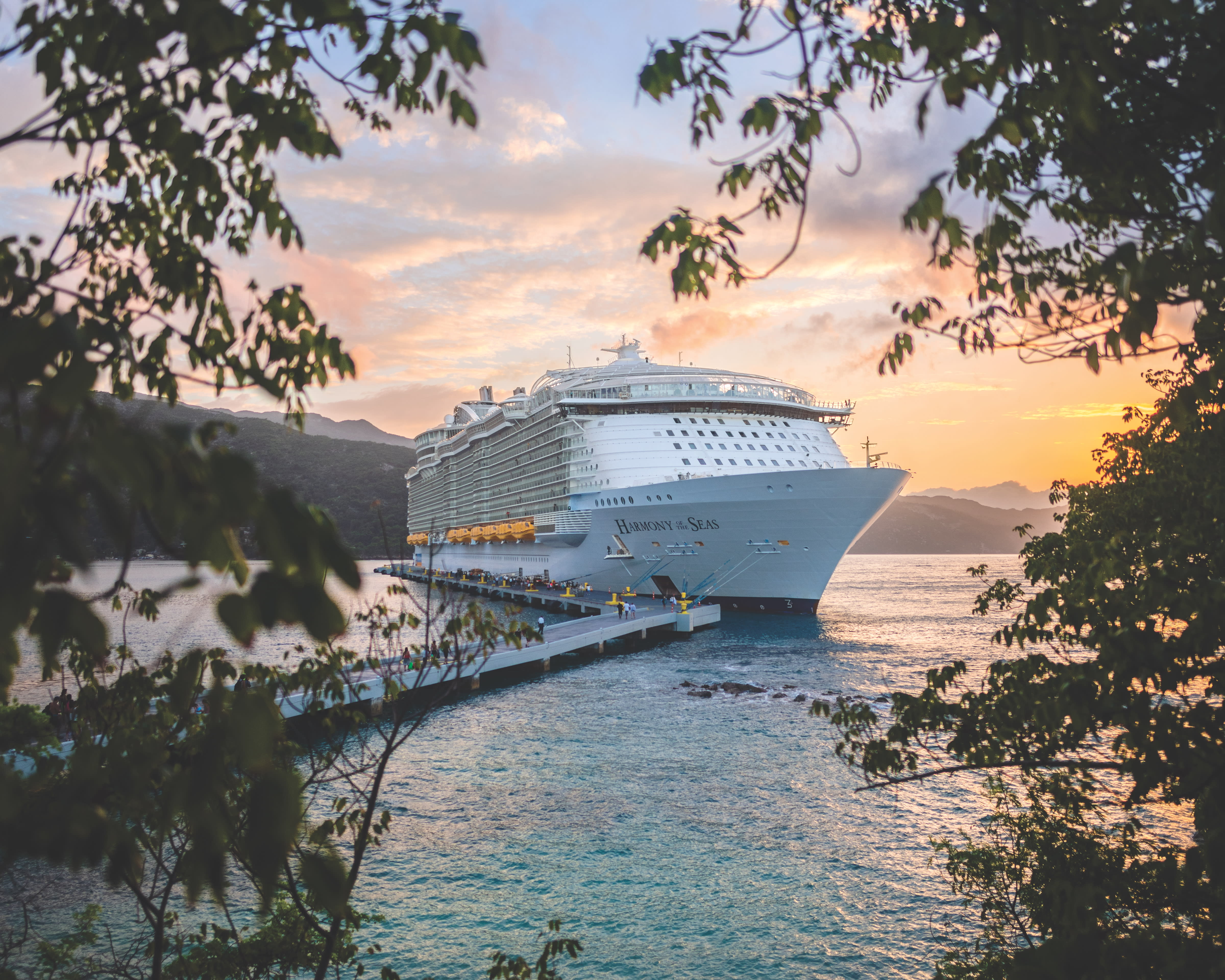 A cruise ship docked off the coast of a tropical island with the sun setting in the background