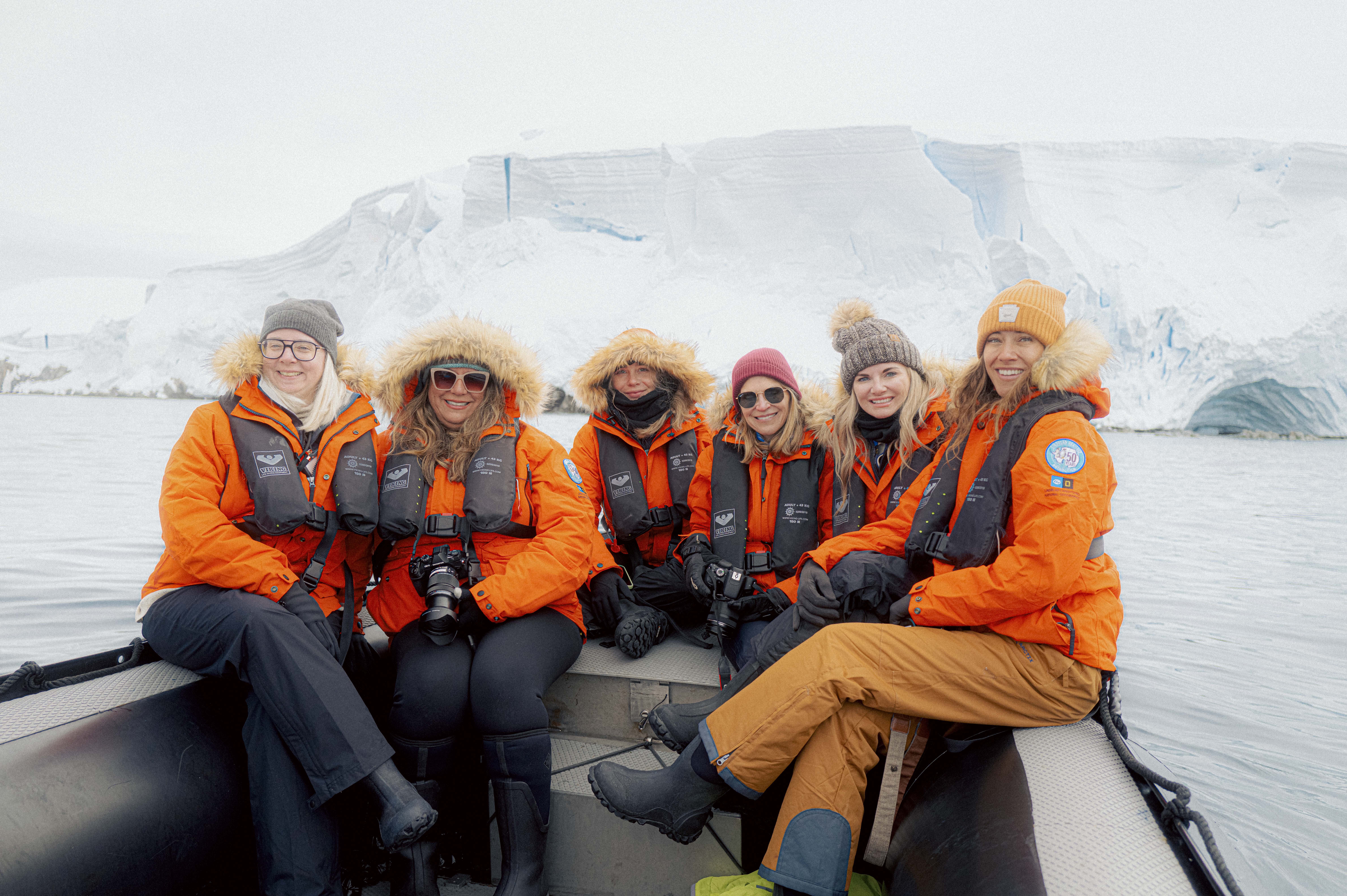 A group of six women dressed in bright orange parkas, life vests, and winter hats sit together on a boat in Antarctica, smiling at the camera with a backdrop of massive icebergs and snowy cliffs.