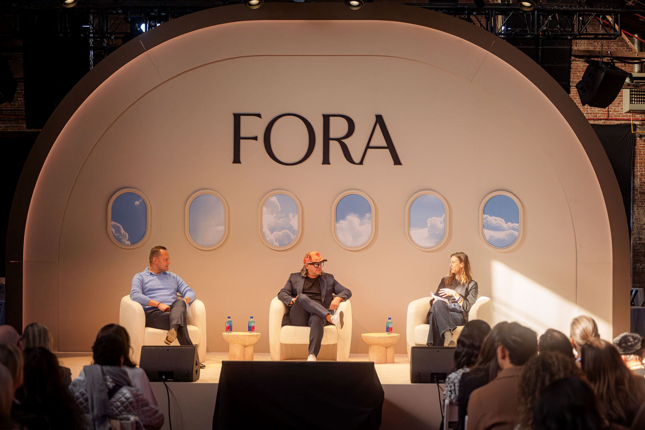 Three speakers on stage at a Fora event, seated in front of a backdrop styled like airplane windows, engaging with an audience.