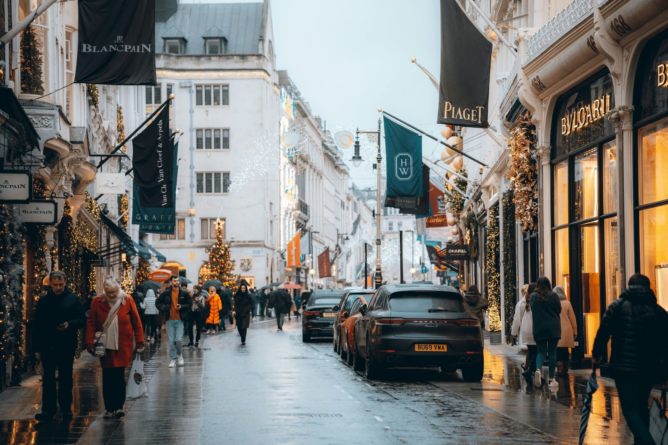 Shoppers and cars line a festive luxury shopping street, with storefronts lit for the holidays and brand banners overhead.