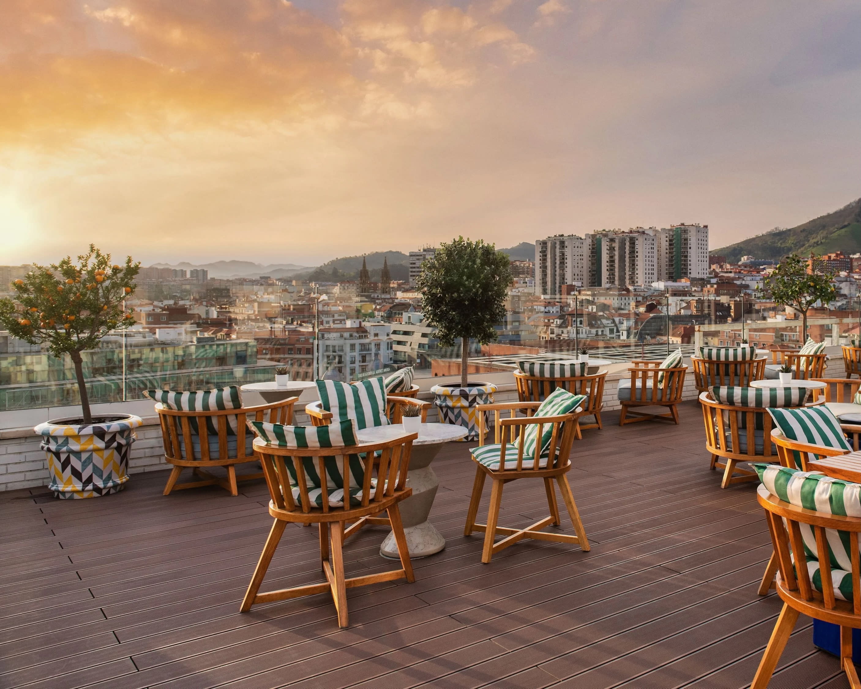 A rooftop terrace with tables and chairs, city in the background, and the sun setting on the horizon