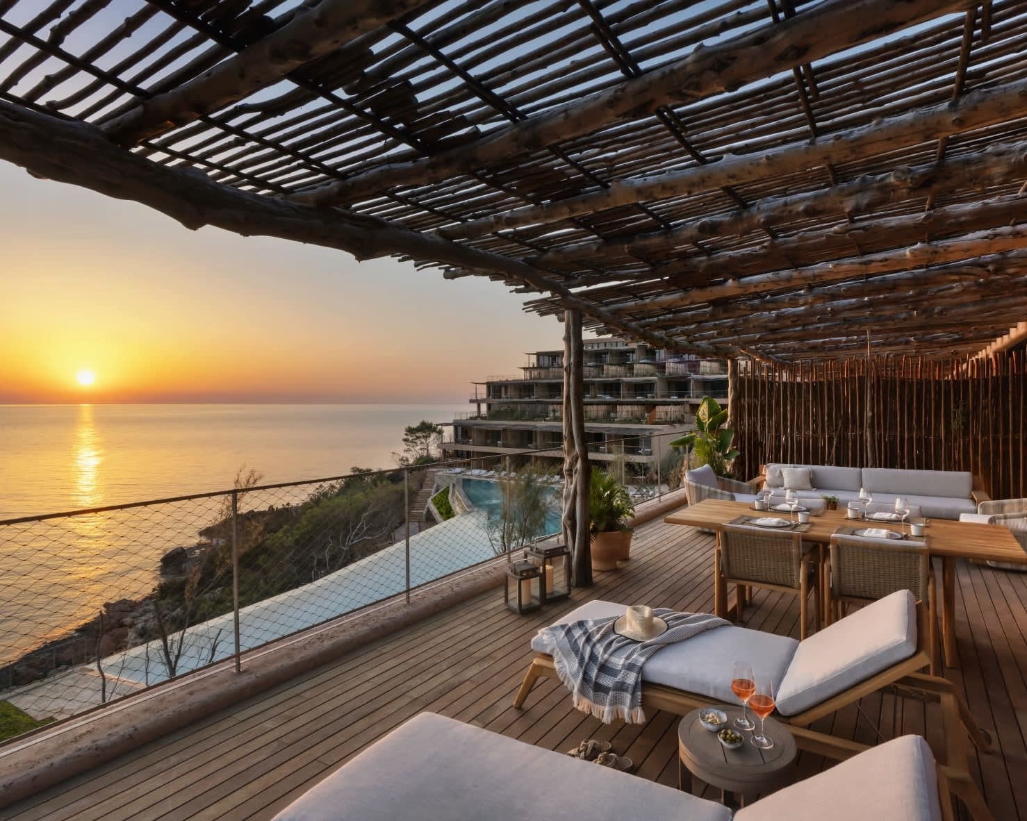 A wooden thatched roof balcony with outdoor furniture overlooking an oceanside resort as the sun sets on the horizon