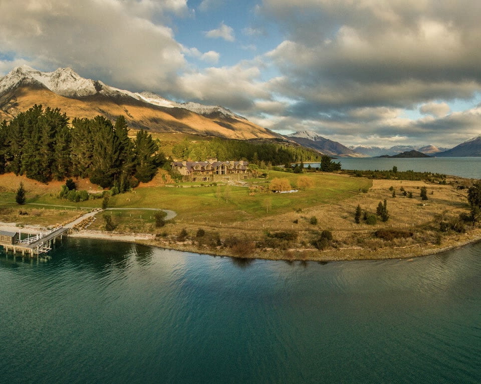 A remote lodge on a peninsula next to a lake with snow capped and green mountains around during day with clouds