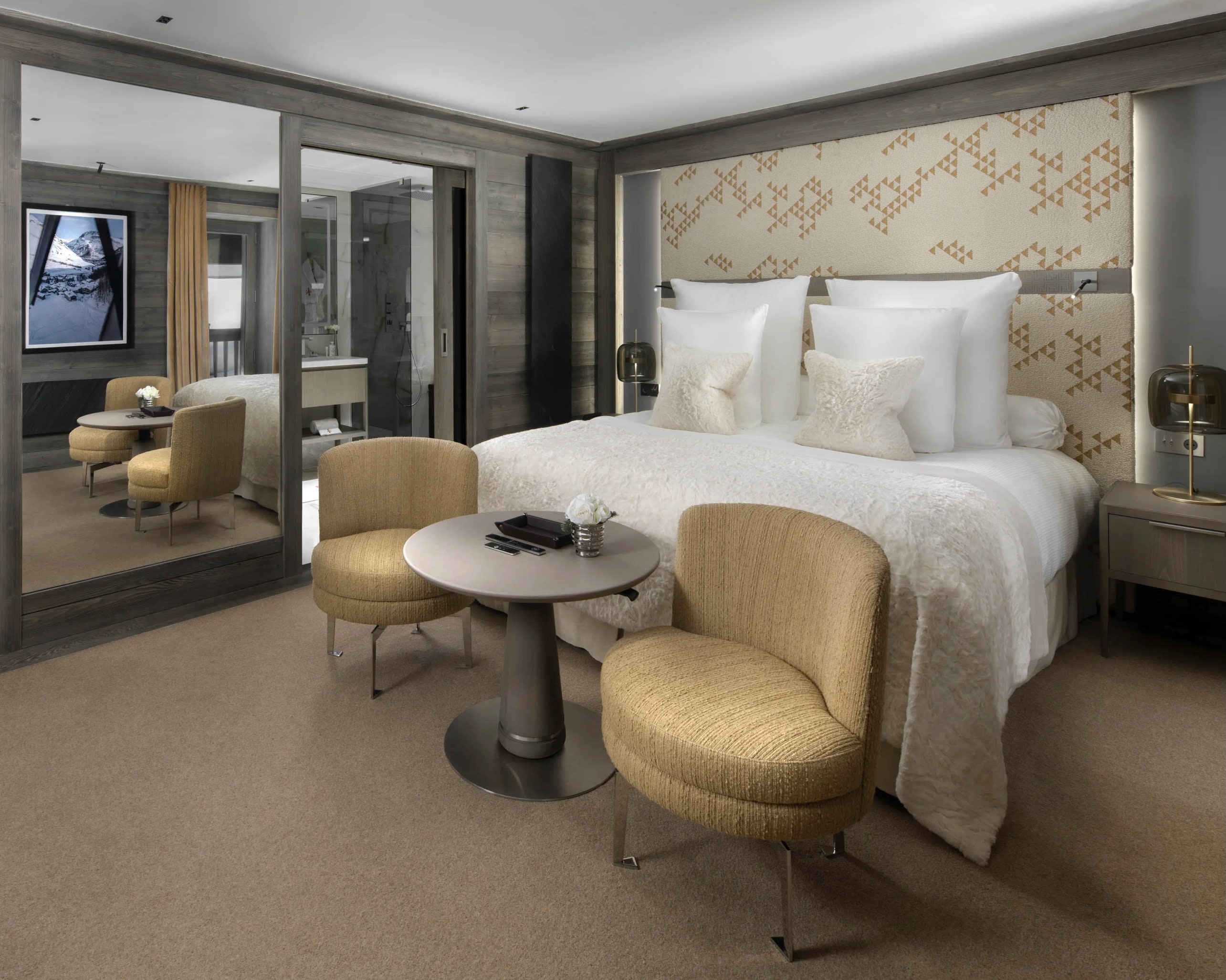A luxurious modern room with white bed linens, gold chairs at foot of the bed, beige headboard, and glass closet next to the bed