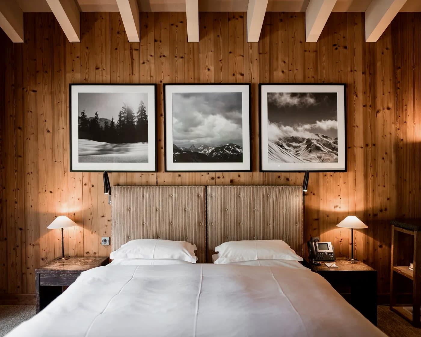 hotel room with white linen bed, wood wall and beam ceiling ith black and white framed photos above the bed