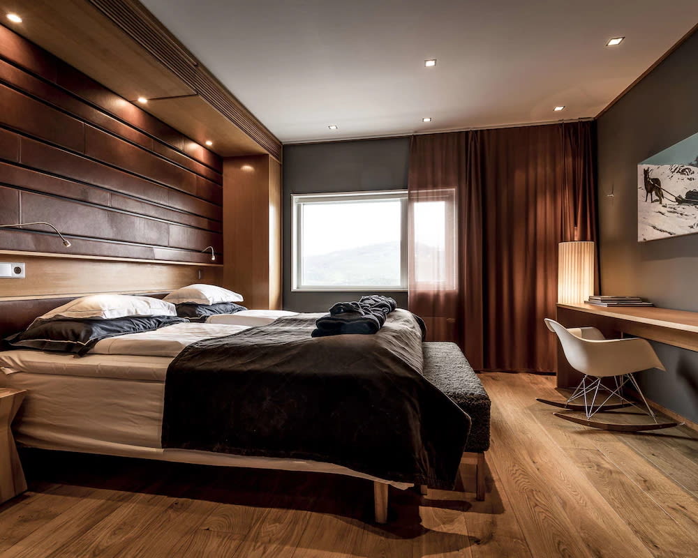 hotel room with white sheets and grey blanket on the bed, wood floors and walls, and a modern white desk chair