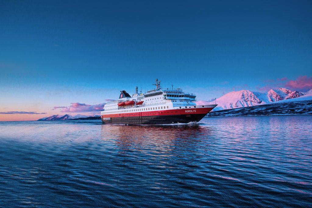 A white, red, and black ship at sea with snow covered mountains at dusk in the background