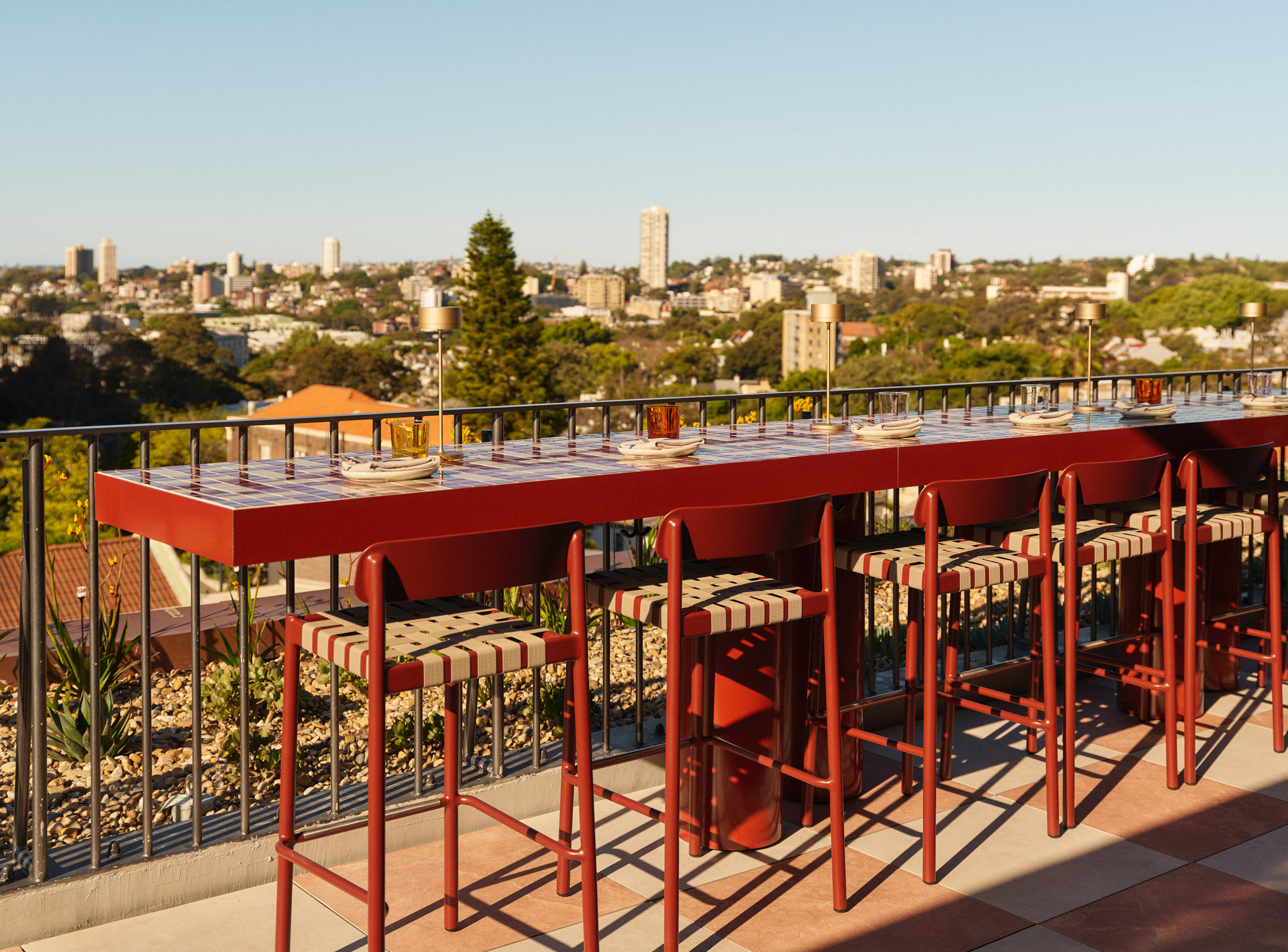 Rooftop bar with red tables and chairs overlooking a city skyline, set for dining under clear blue skies with warm afternoon light.