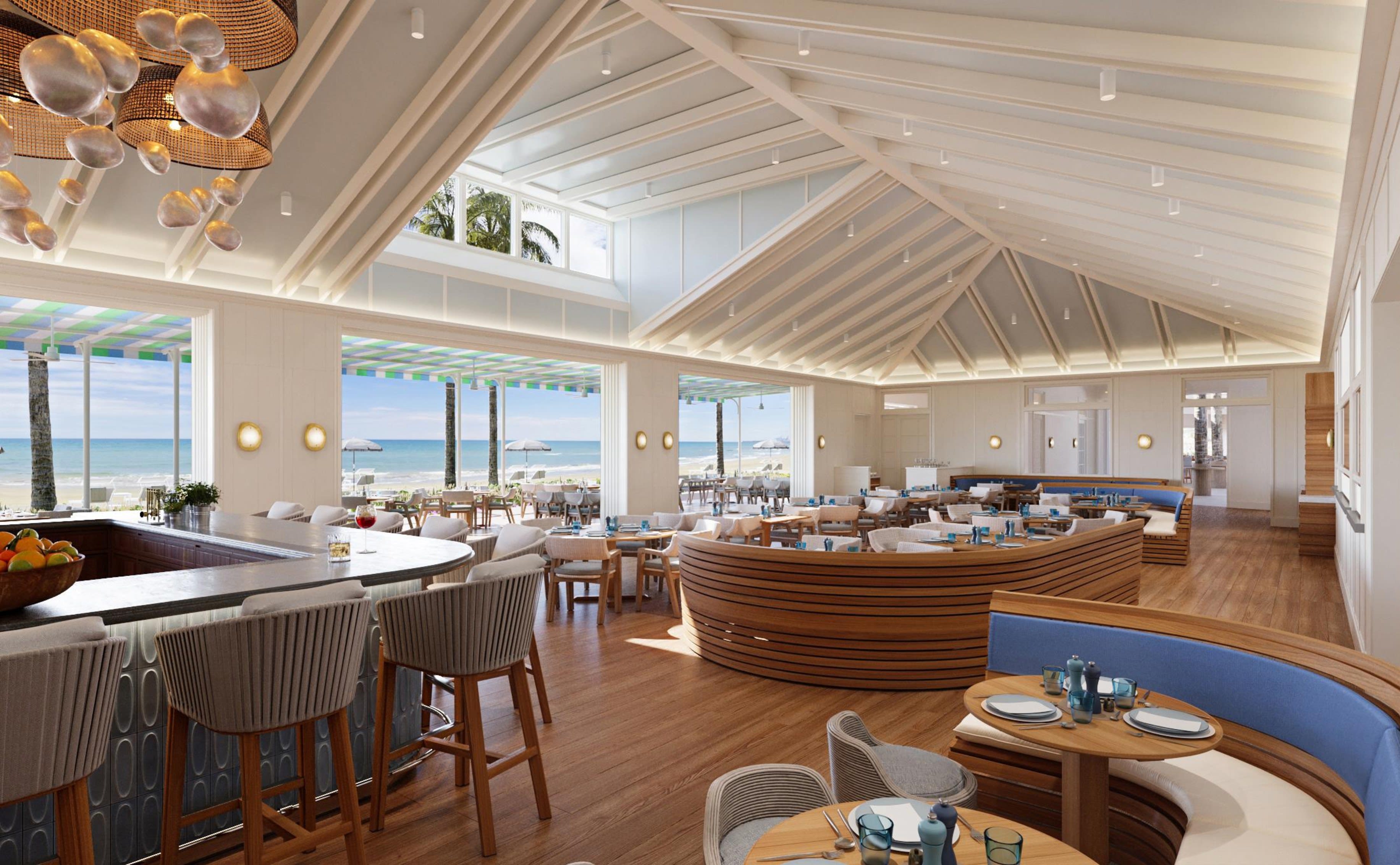 Bright beachfront restaurant with vaulted ceilings, curved wooden seating, and ocean views, featuring a relaxed coastal design and open-air dining area.